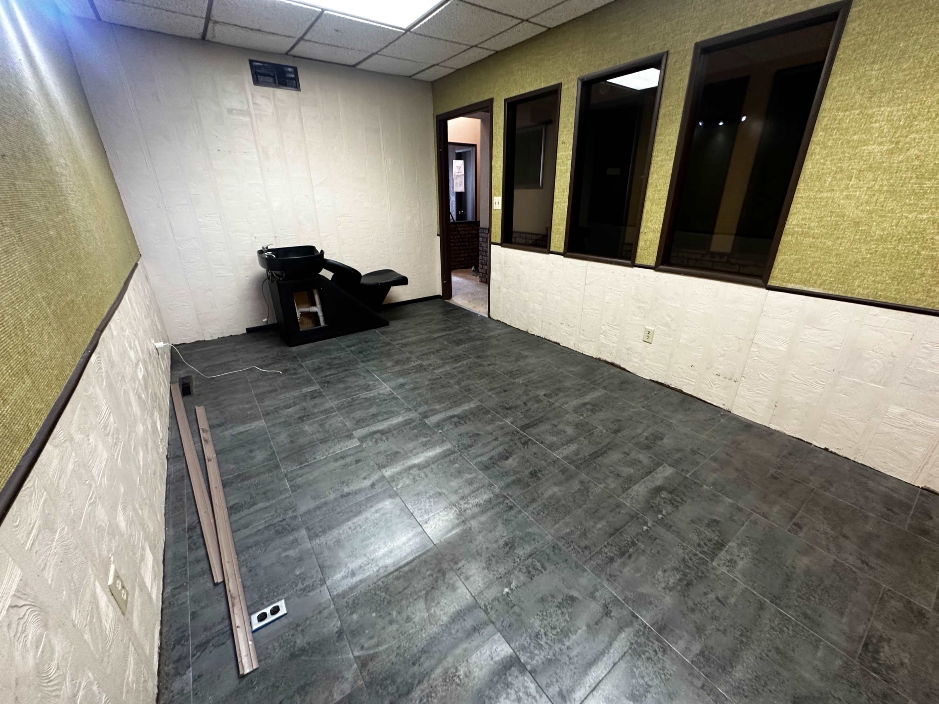 The image depicts an empty room with gray tiled flooring, a few windows along the walls, and a black chair positioned in one corner.