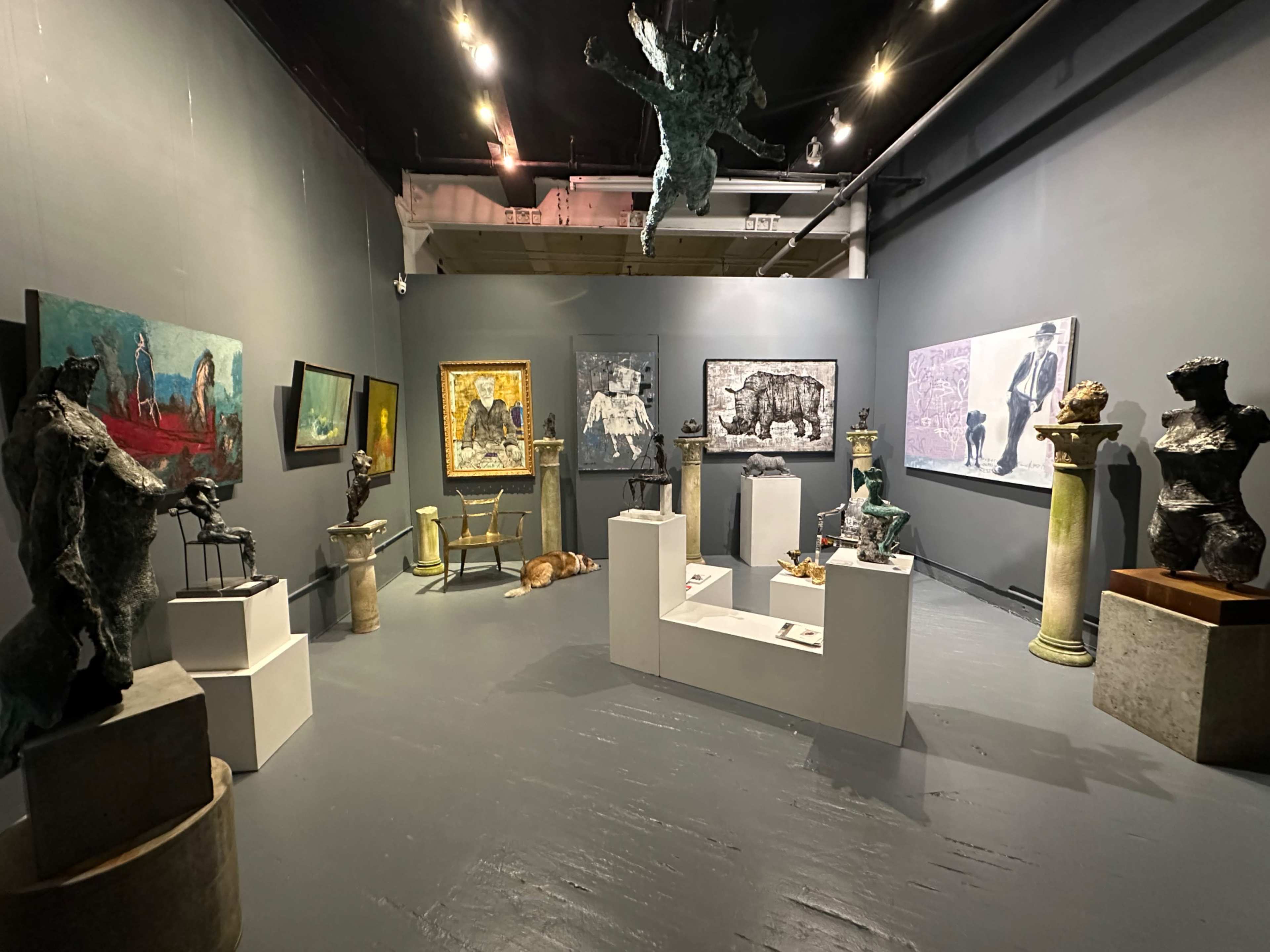 The image shows an art gallery featuring various sculptures and paintings displayed on gray walls and a gray floor, with a dog lying on the floor.
