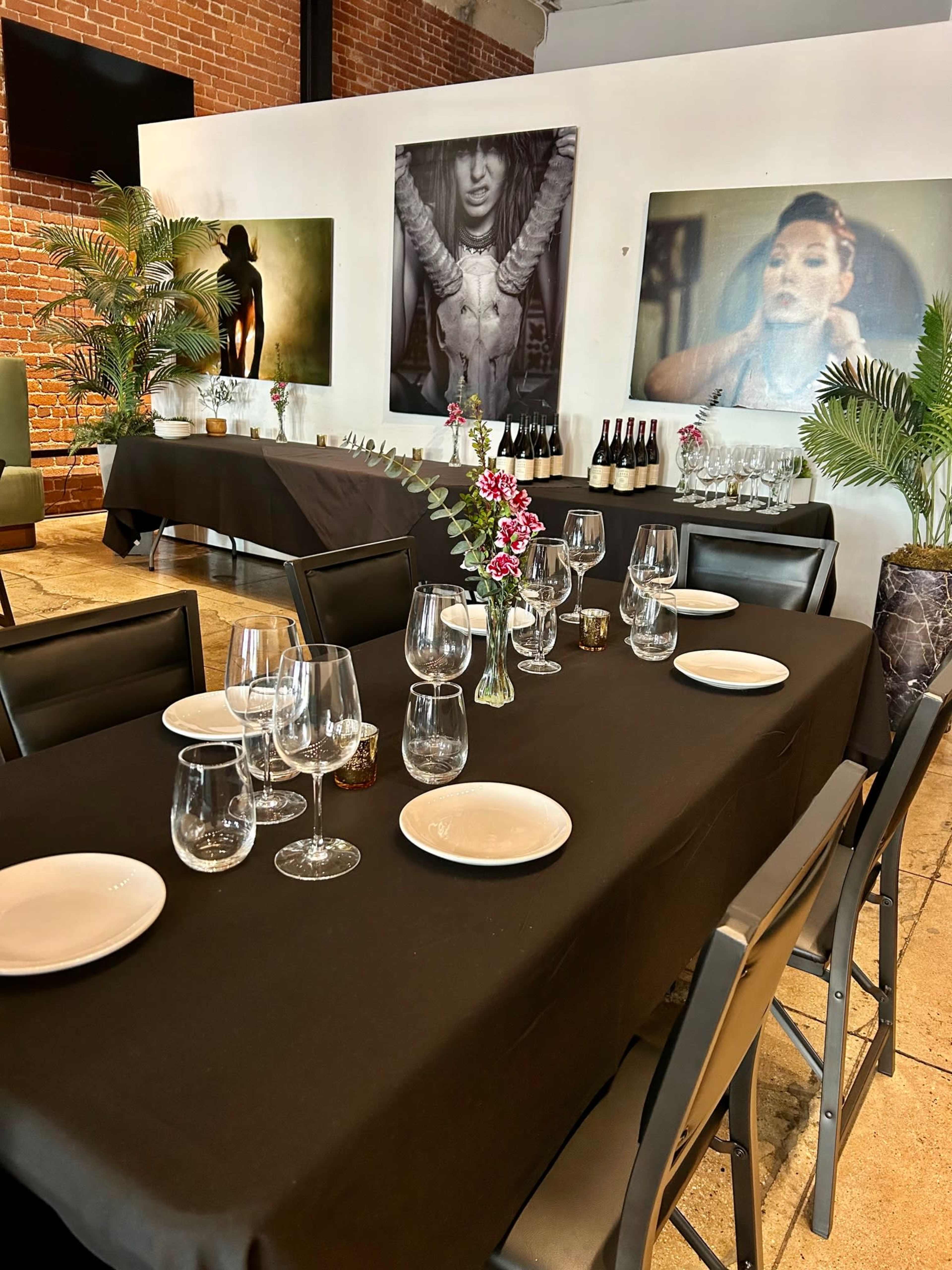 The image shows a dining setup with black tablecloths, empty glassware, and white plates arranged on tables, surrounded by decorative plants and wall art.