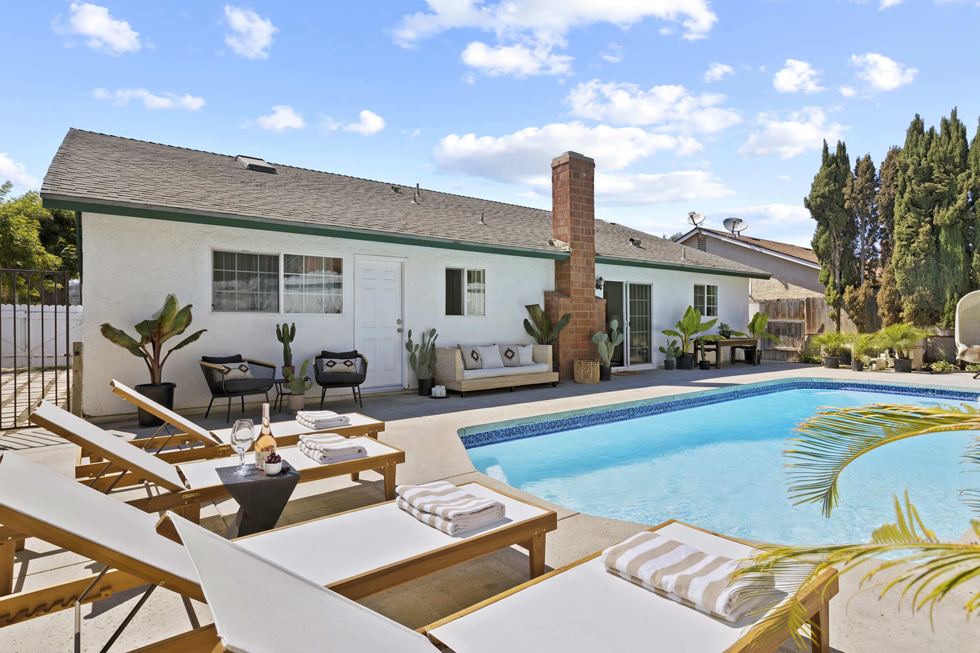 The image shows a backyard scene featuring a swimming pool surrounded by lounge chairs, a small table with drinks, and a house in the background with a patio area.