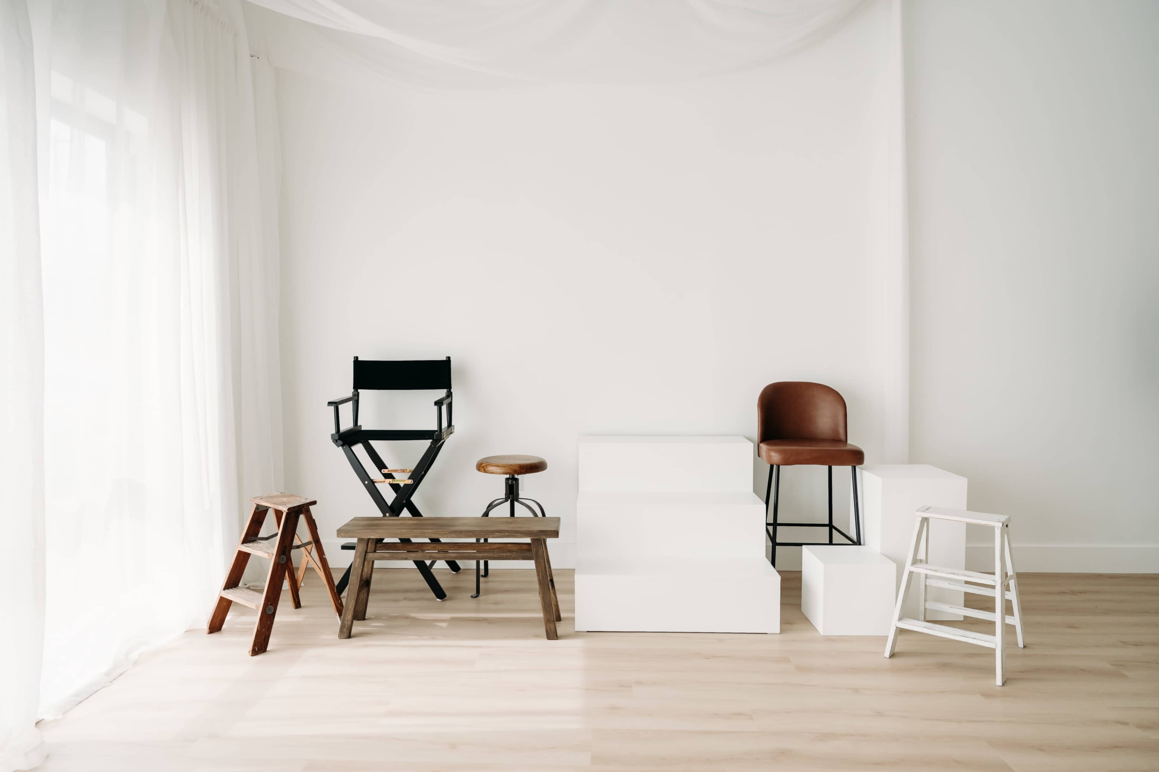 The image shows a minimalist interior space with various types of chairs and stools arranged against a white wall.