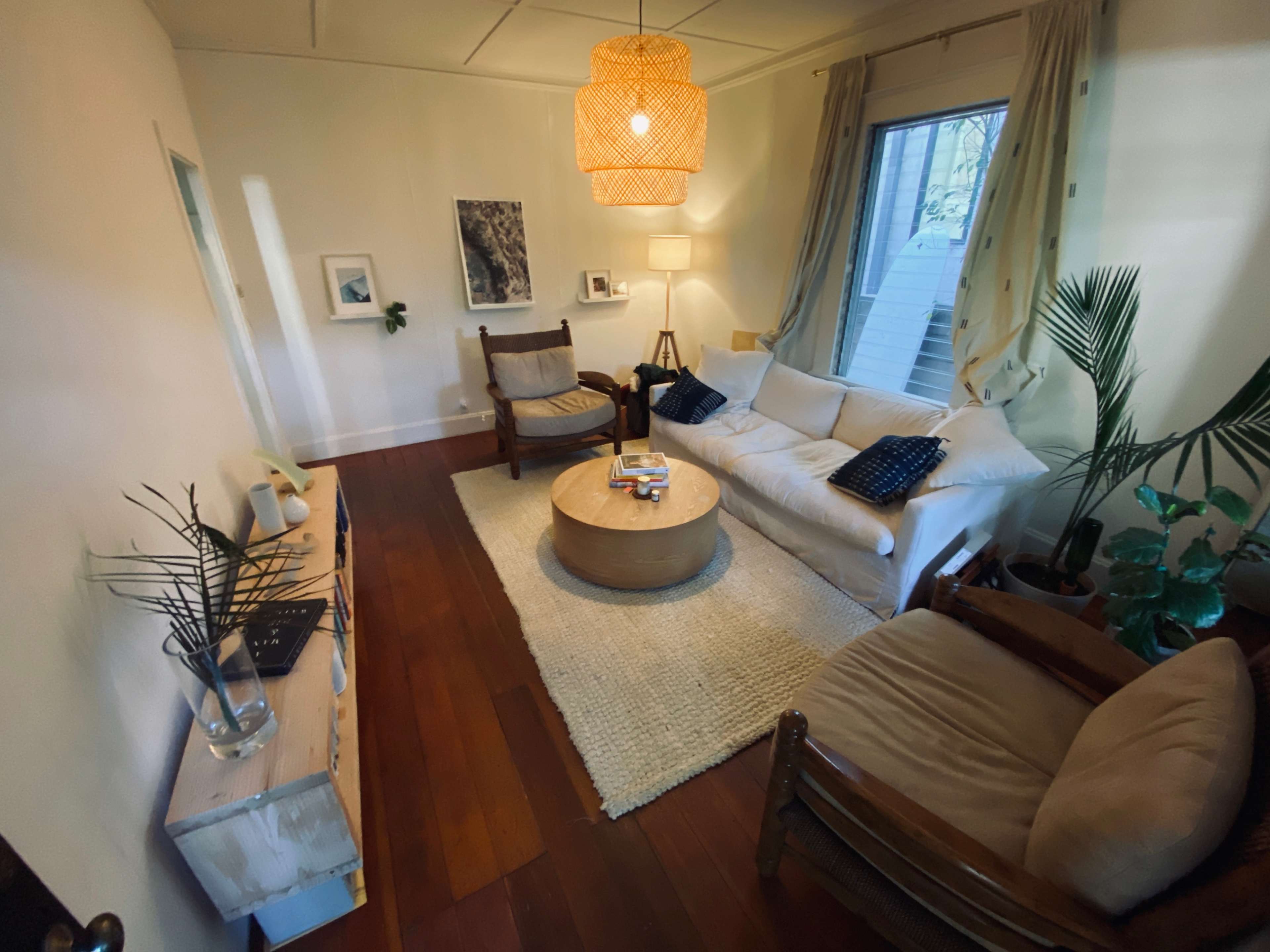 The image shows a cozy living room with a light-colored sofa, wooden furniture, and a round coffee table on a woven rug, surrounded by plants and framed artwork.