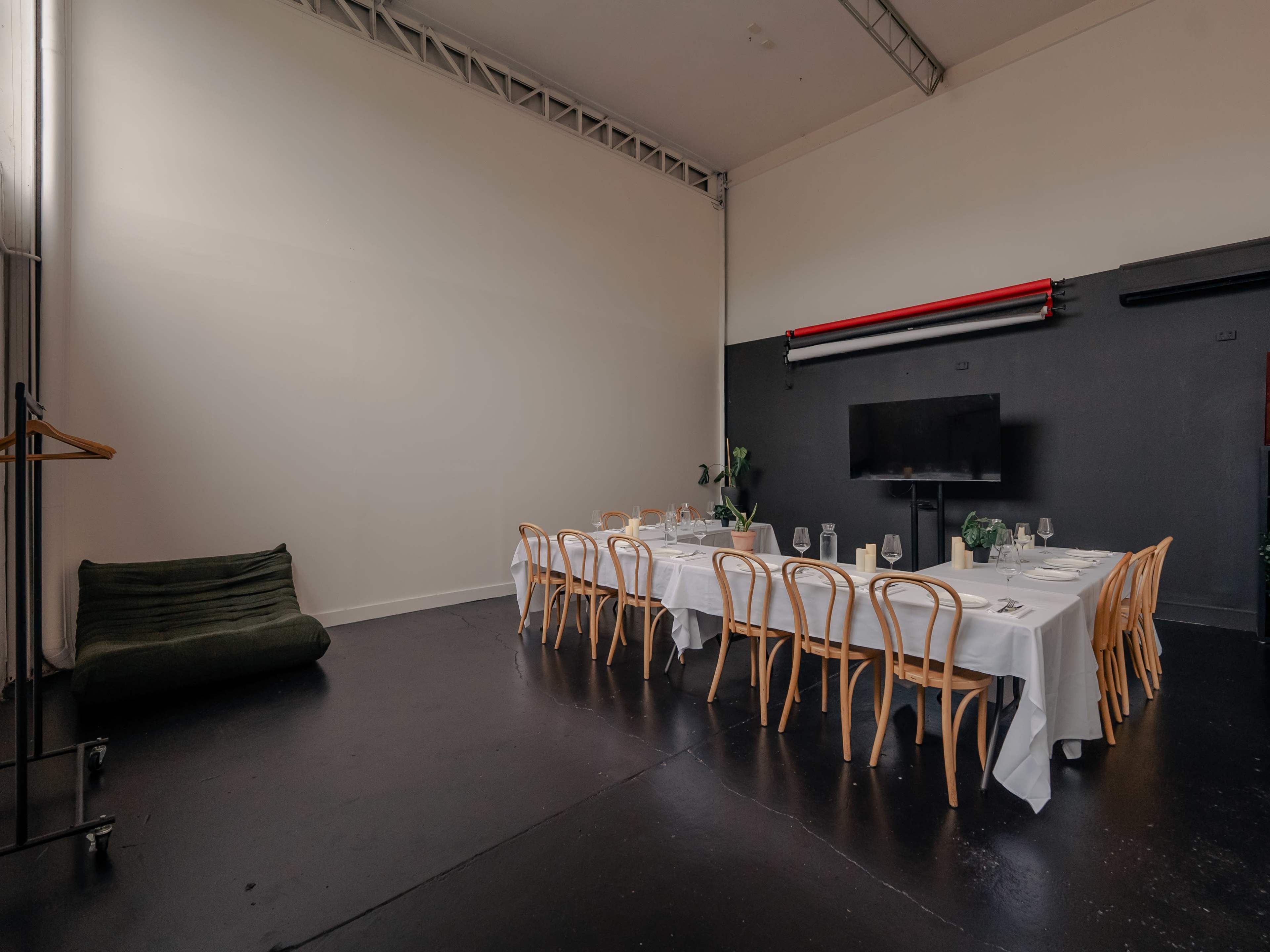 A long dining table with wooden chairs is set in a minimally decorated room with dark flooring and a flat-screen television on the wall.