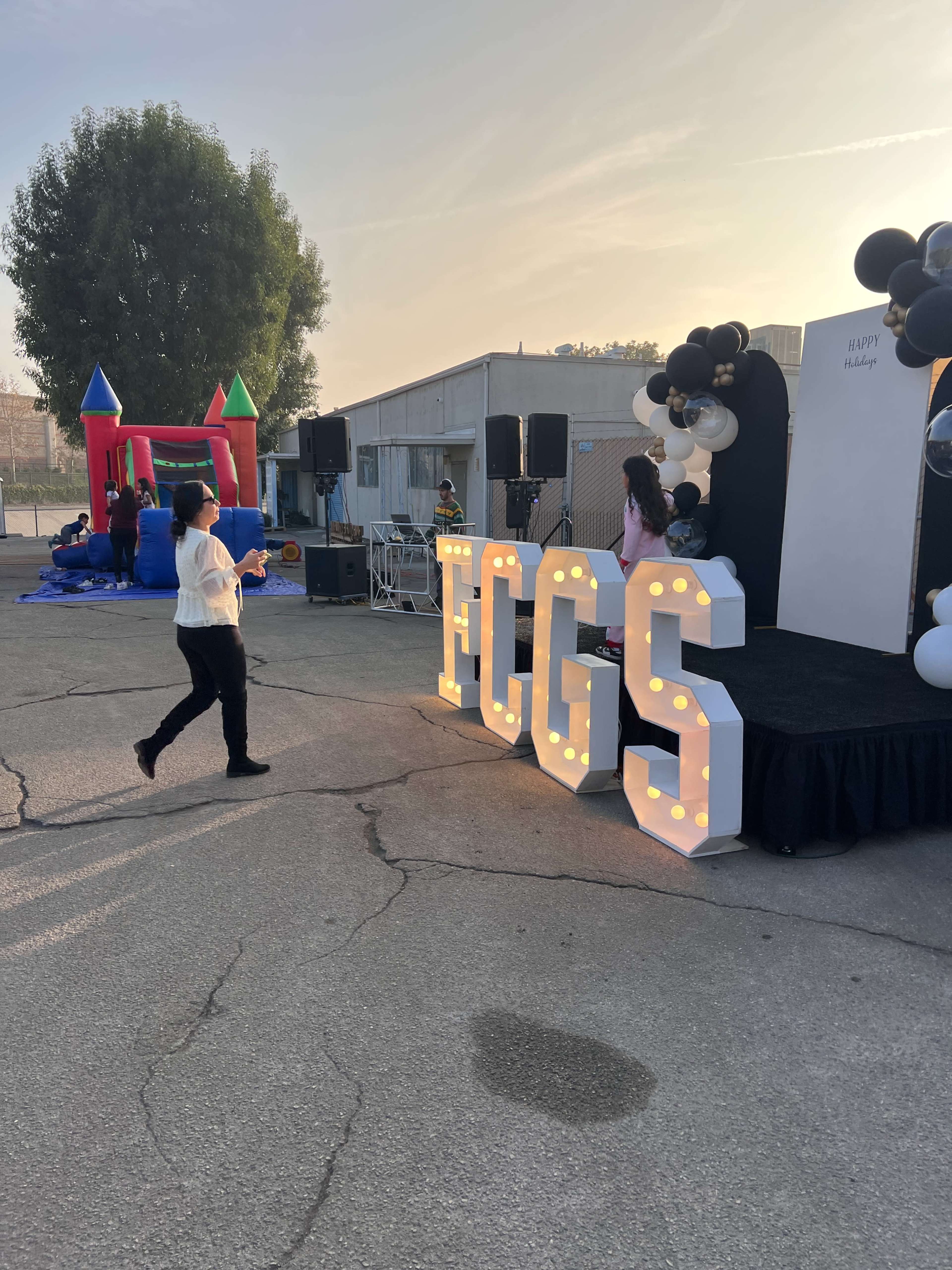 A person walks toward a decorated stage with illuminated letters spelling "FCGS" at an outdoor event, while colorful inflatable structures are visible in the background.