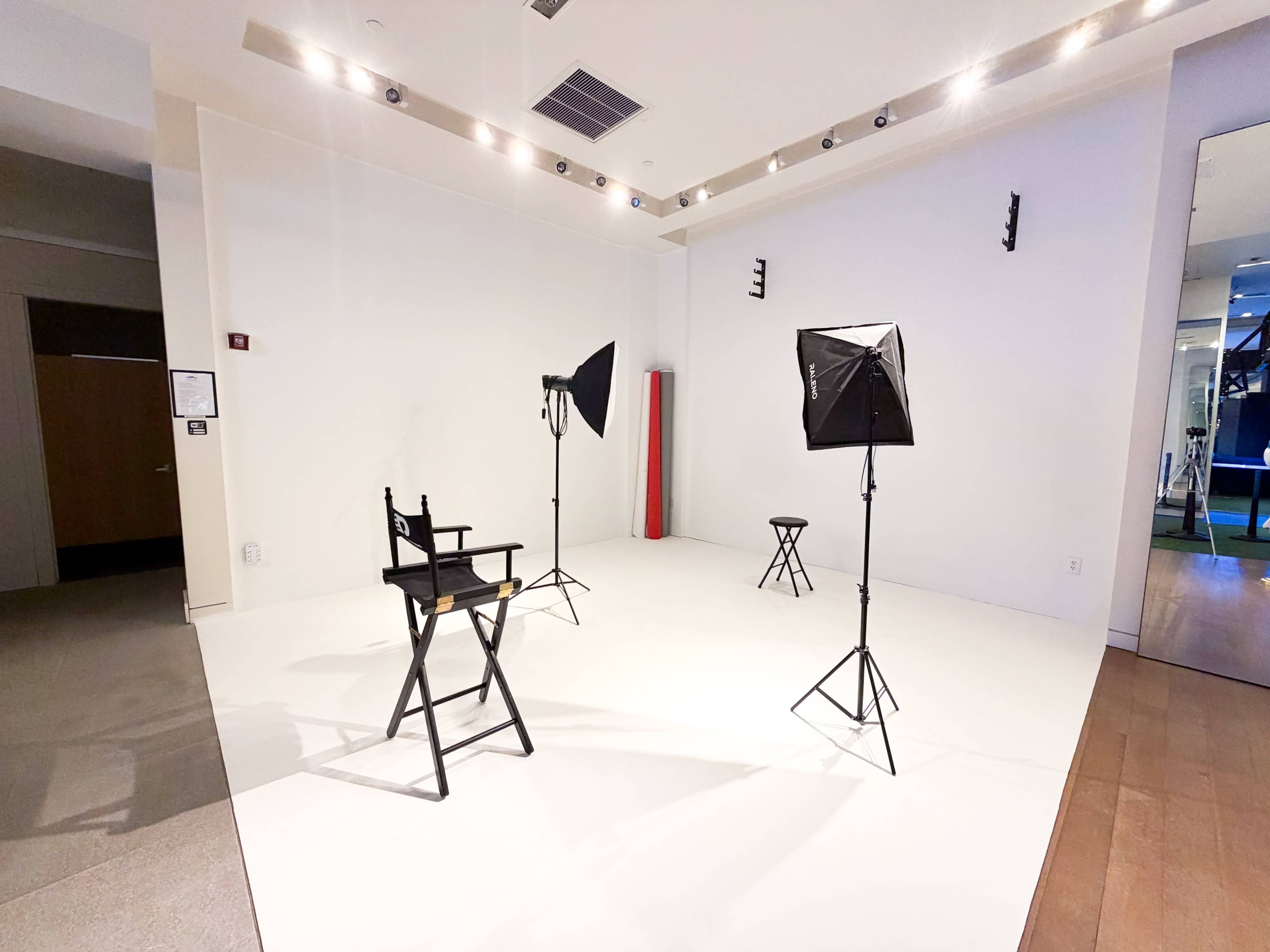The image shows a minimalist studio space with three light stands, a director's chair, and a small stool arranged on a white floor.