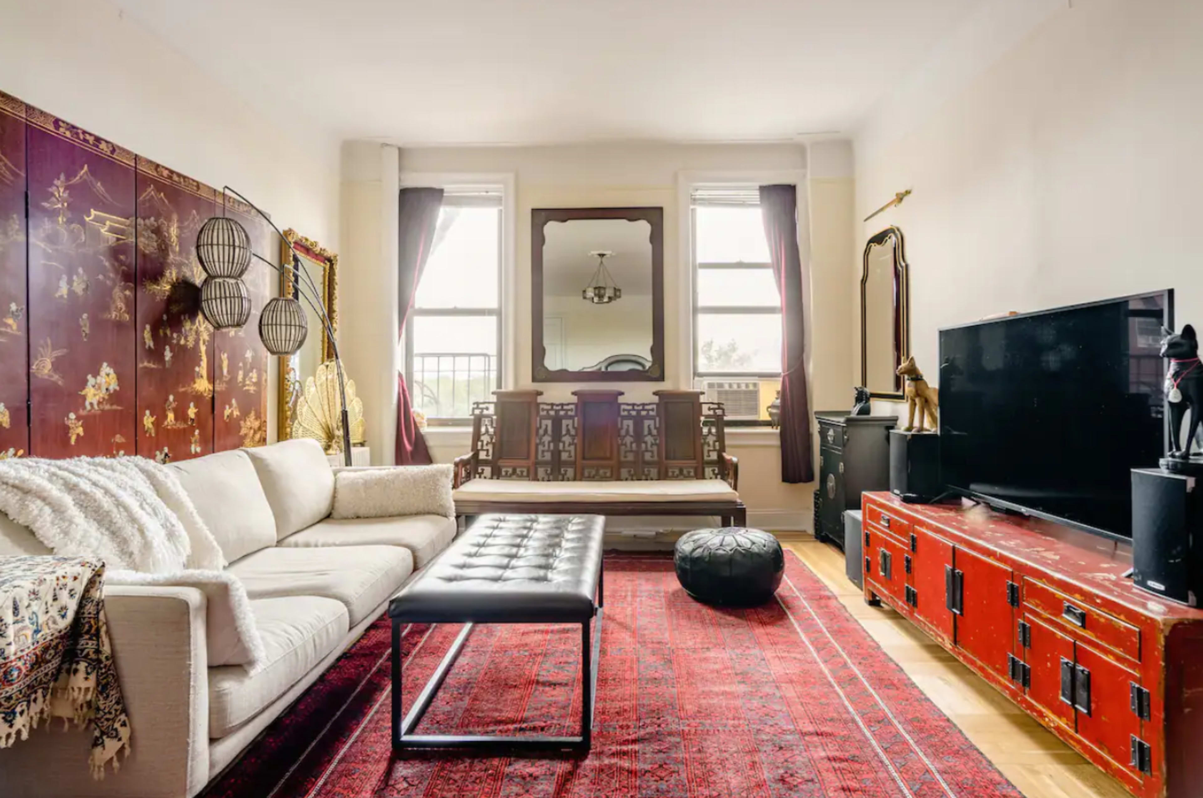 The living room features a white sofa, a black leather ottoman, a red area rug, and decorative elements including a large mirrored wall and a red storage cabinet.