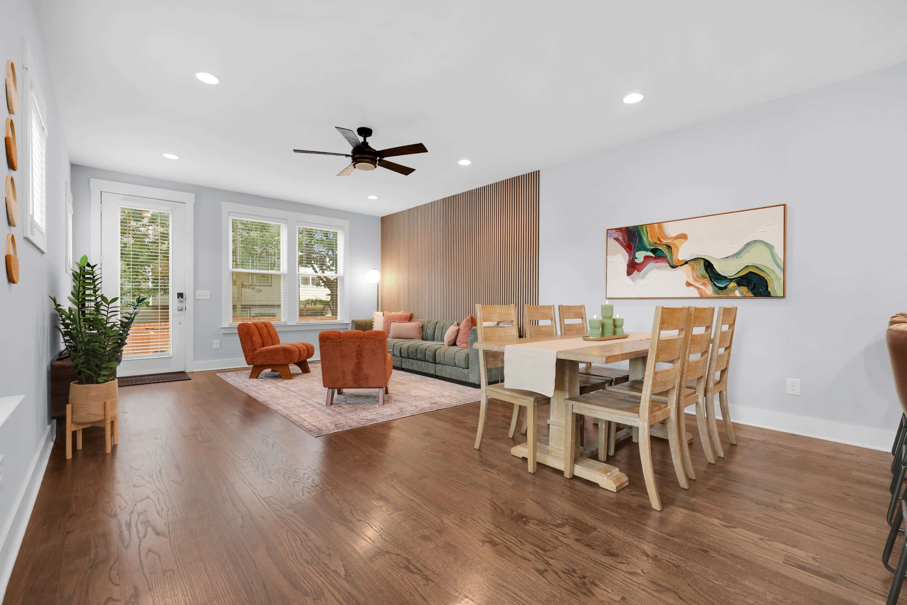 The image shows a well-lit living and dining area featuring a green sofa, orange armchair, wooden dining table, and large windows.