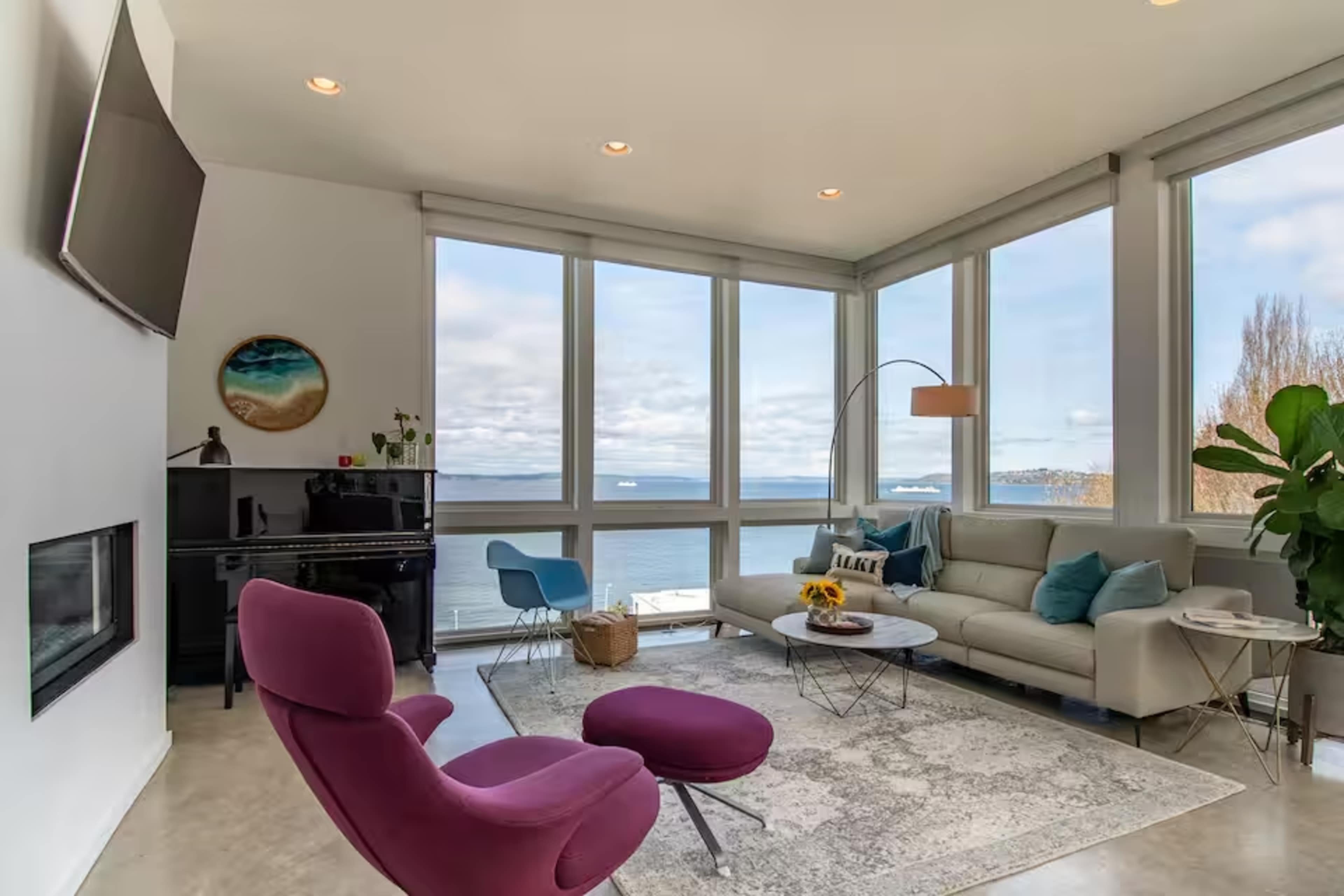A modern living room features large windows overlooking a waterfront view, a gray sofa, a purple chair, a piano, and a round coffee table.