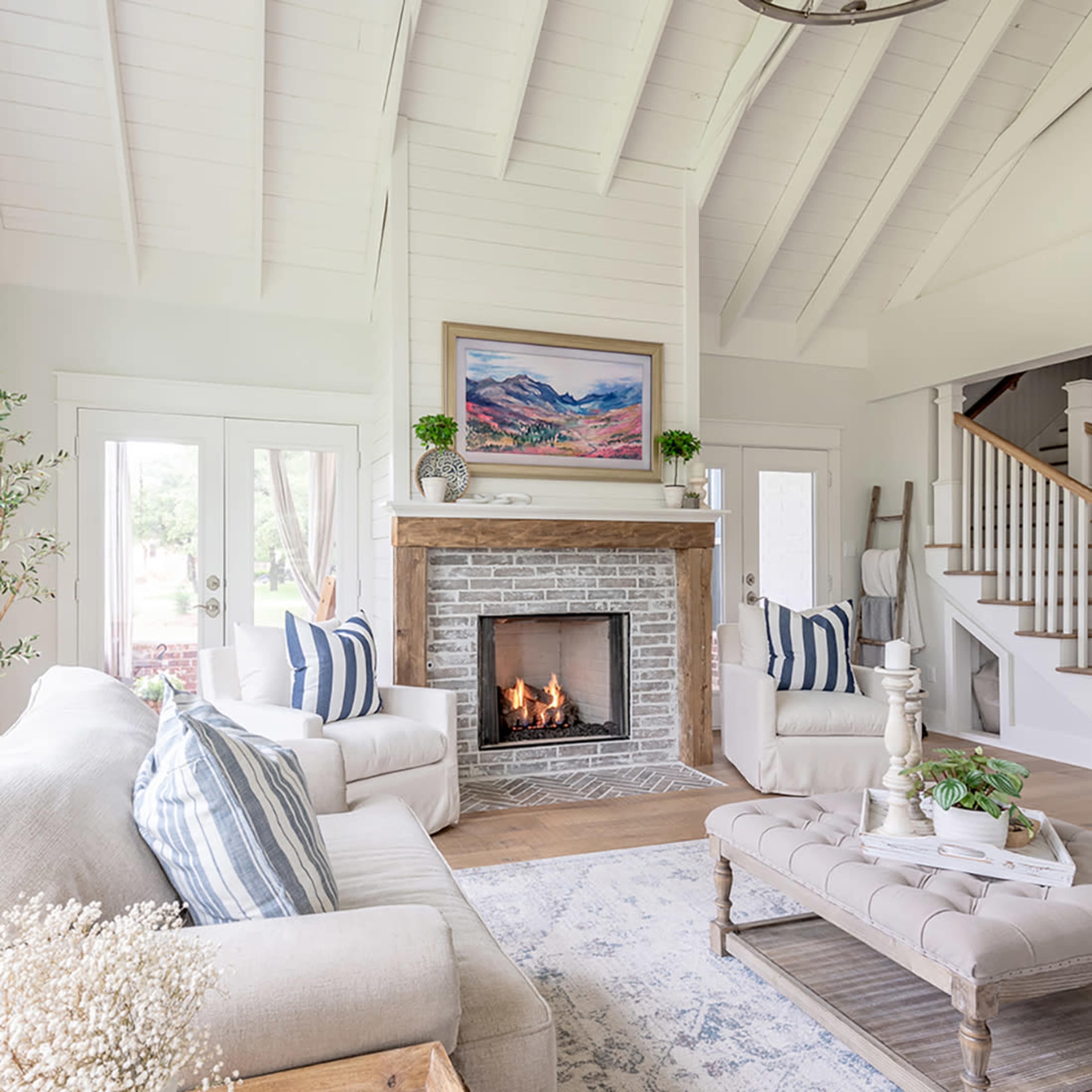 A spacious living room features a brick fireplace with a wooden mantel, surrounded by light-colored furniture and decorative plants.