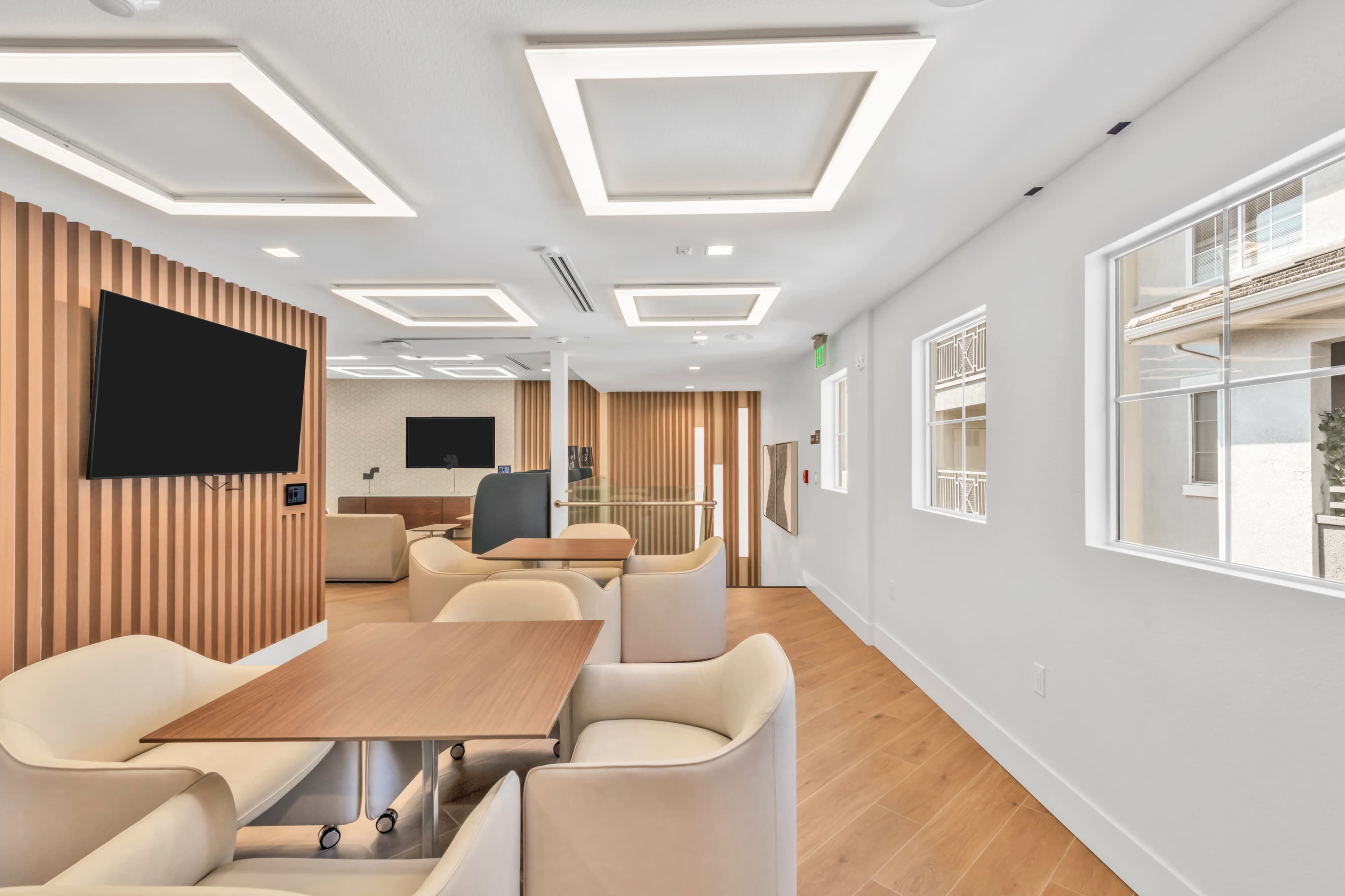 The image shows a modern, well-lit lounge area with light-colored furniture and wooden accents, featuring a large wall-mounted TV and a dining table.