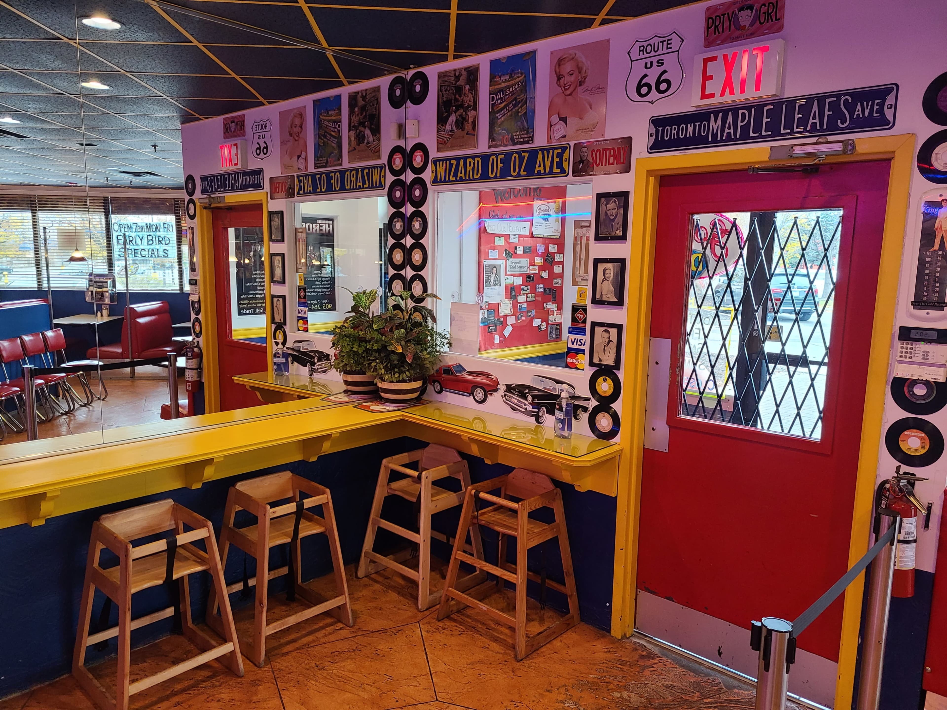 The image shows a brightly colored diner interior featuring a yellow bar with wooden stools, decorative vinyl records on the walls, and a red exit door next to various memorabilia.