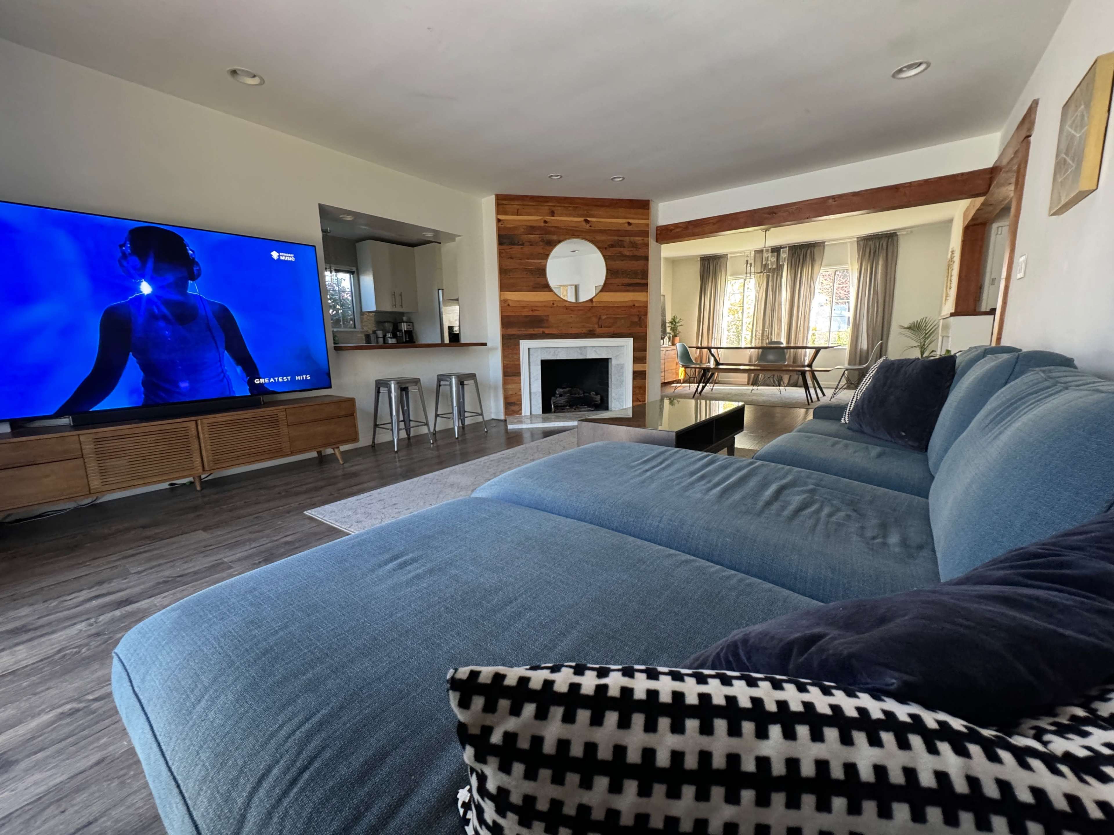 A modern living room featuring a large blue sectional sofa, a wall-mounted television displaying a figure, and an open kitchen area with a dining space in the background.