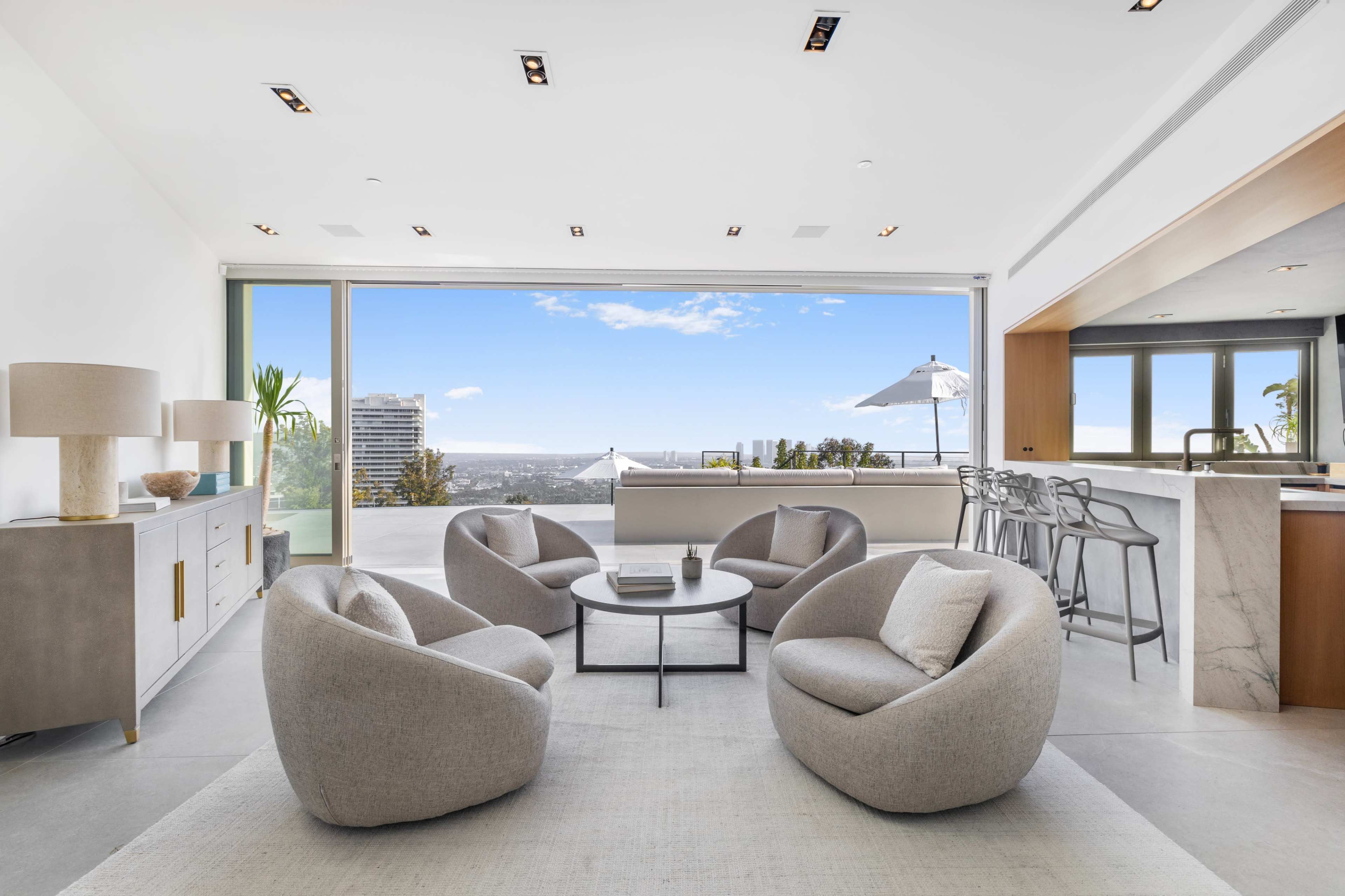 A modern living room features four rounded chairs around a circular table, with large windows offering a view of the city and a kitchen bar in the background.