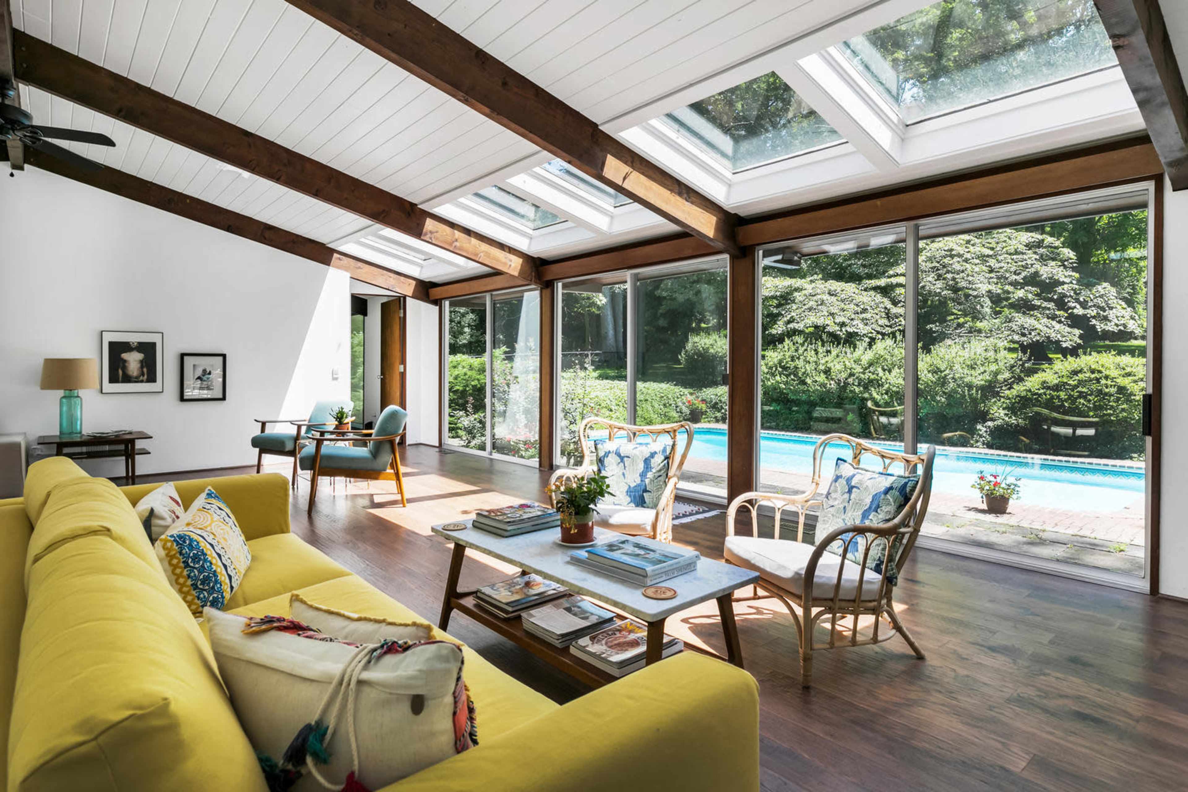 A sunlit living room features a yellow sofa, wooden beams, large glass windows, and a view of a pool surrounded by greenery.