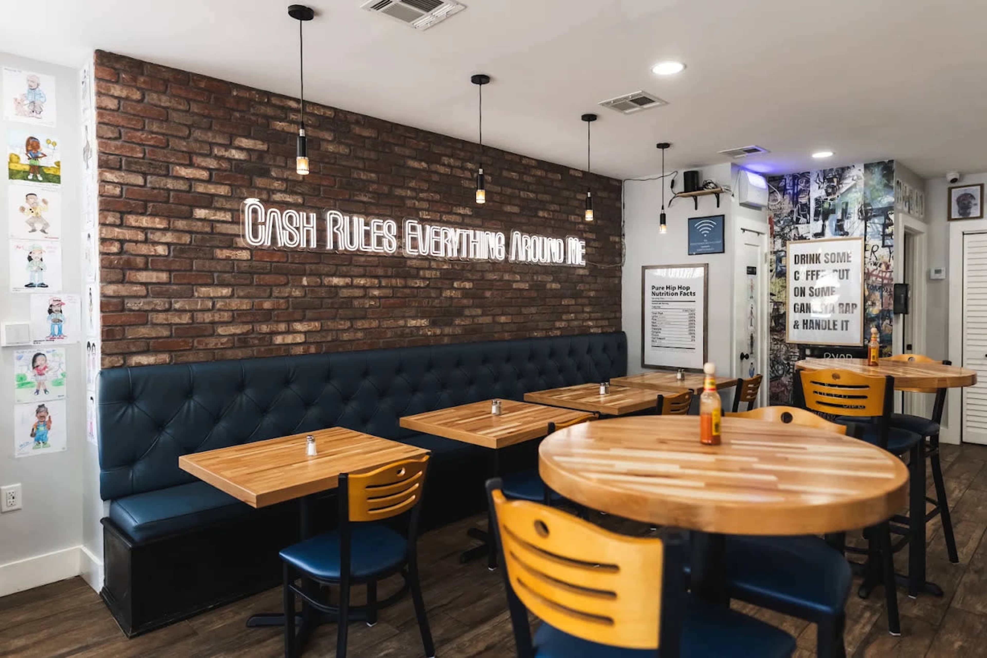 The image shows a modern dining area with wooden tables and blue cushioned seating against a brick wall, featuring hanging light fixtures and a neon sign.