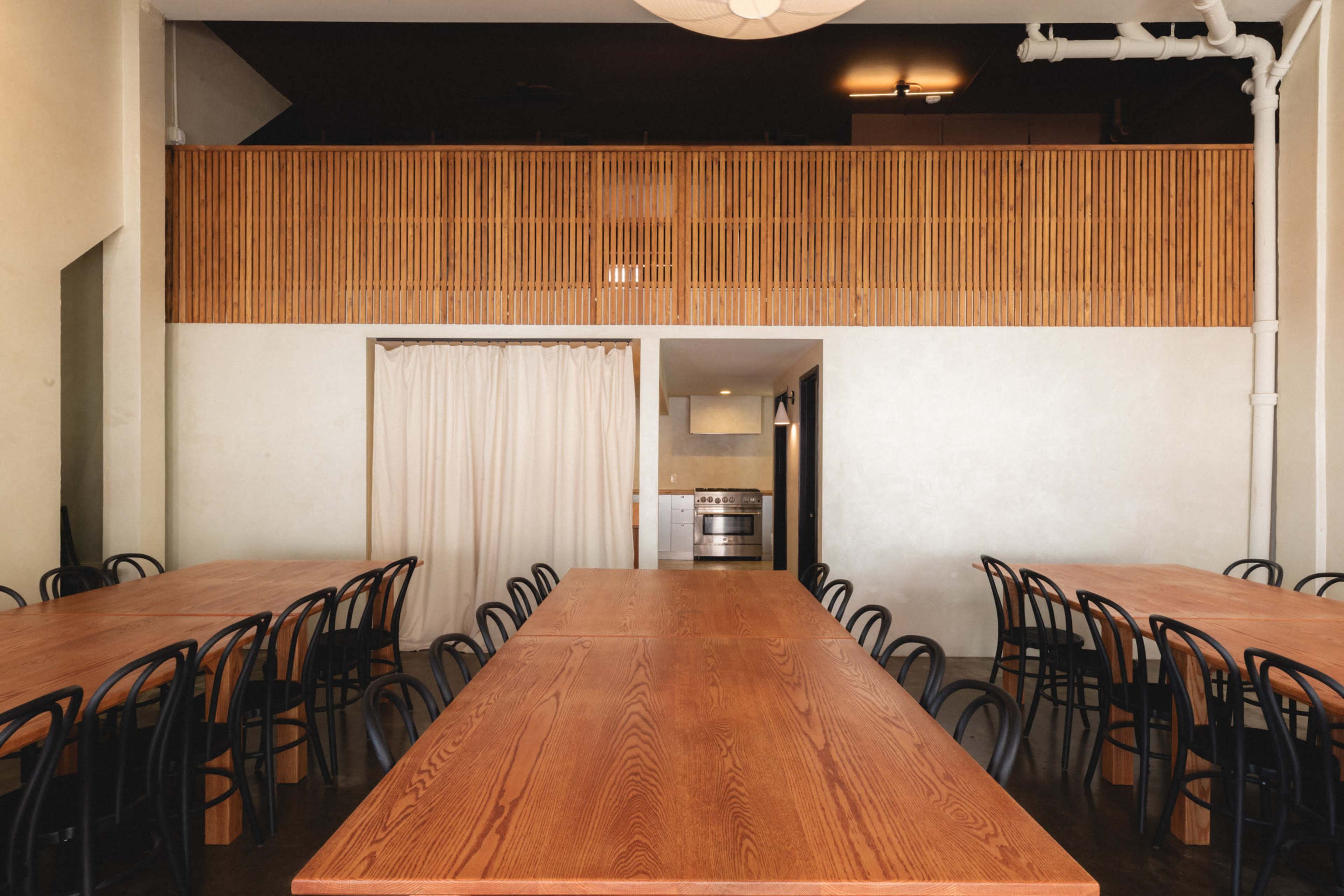 The image shows a spacious room with multiple large wooden tables arranged linearly and a kitchen area visible in the background behind a curtain.