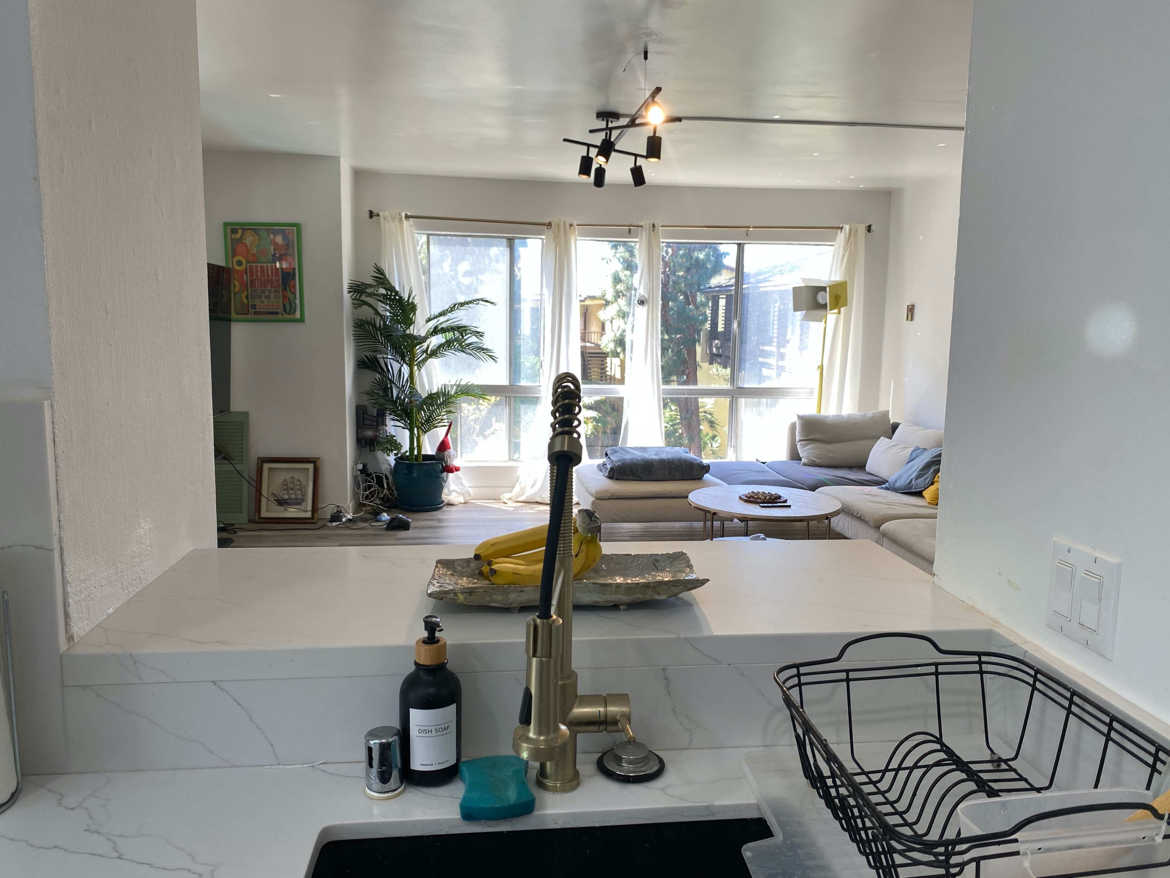 A kitchen with a modern faucet and sink overlooks a living room featuring a large sofa, plants, and natural light streaming through windows.