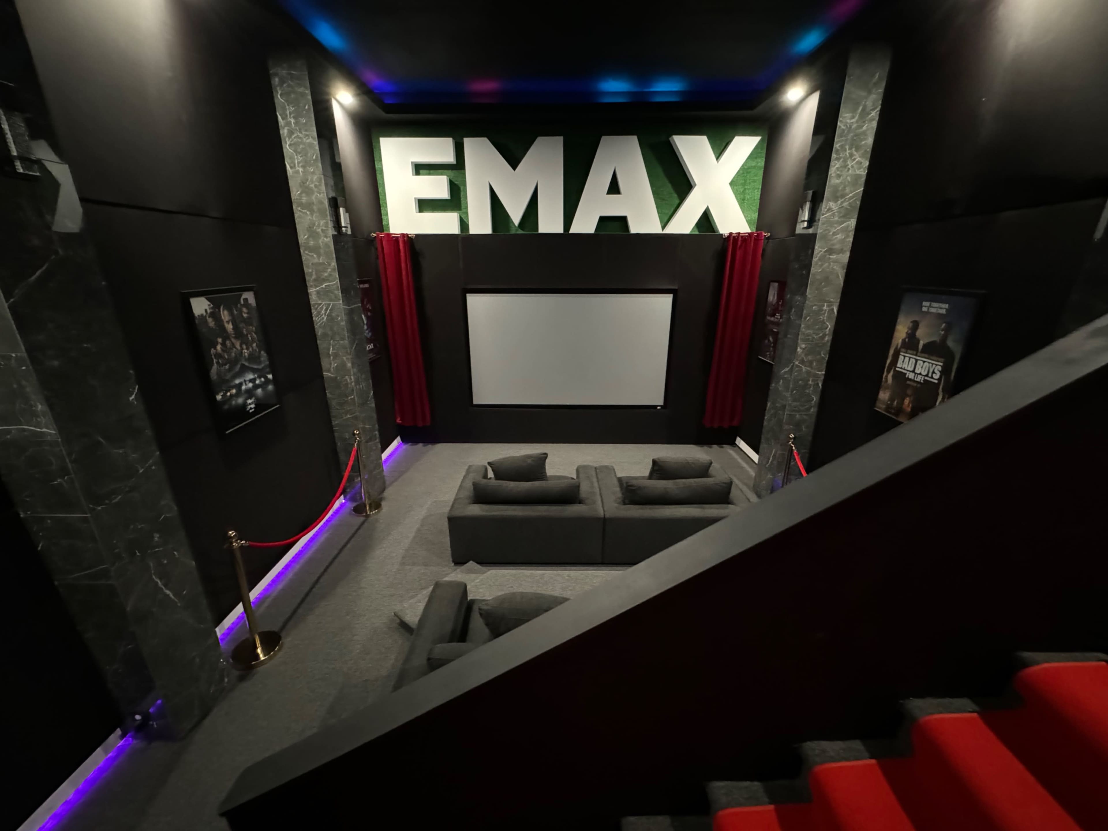 The image shows a movie theater with a large screen, gray sofas arranged in front, and prominent signage reading "EMAX" on the wall.