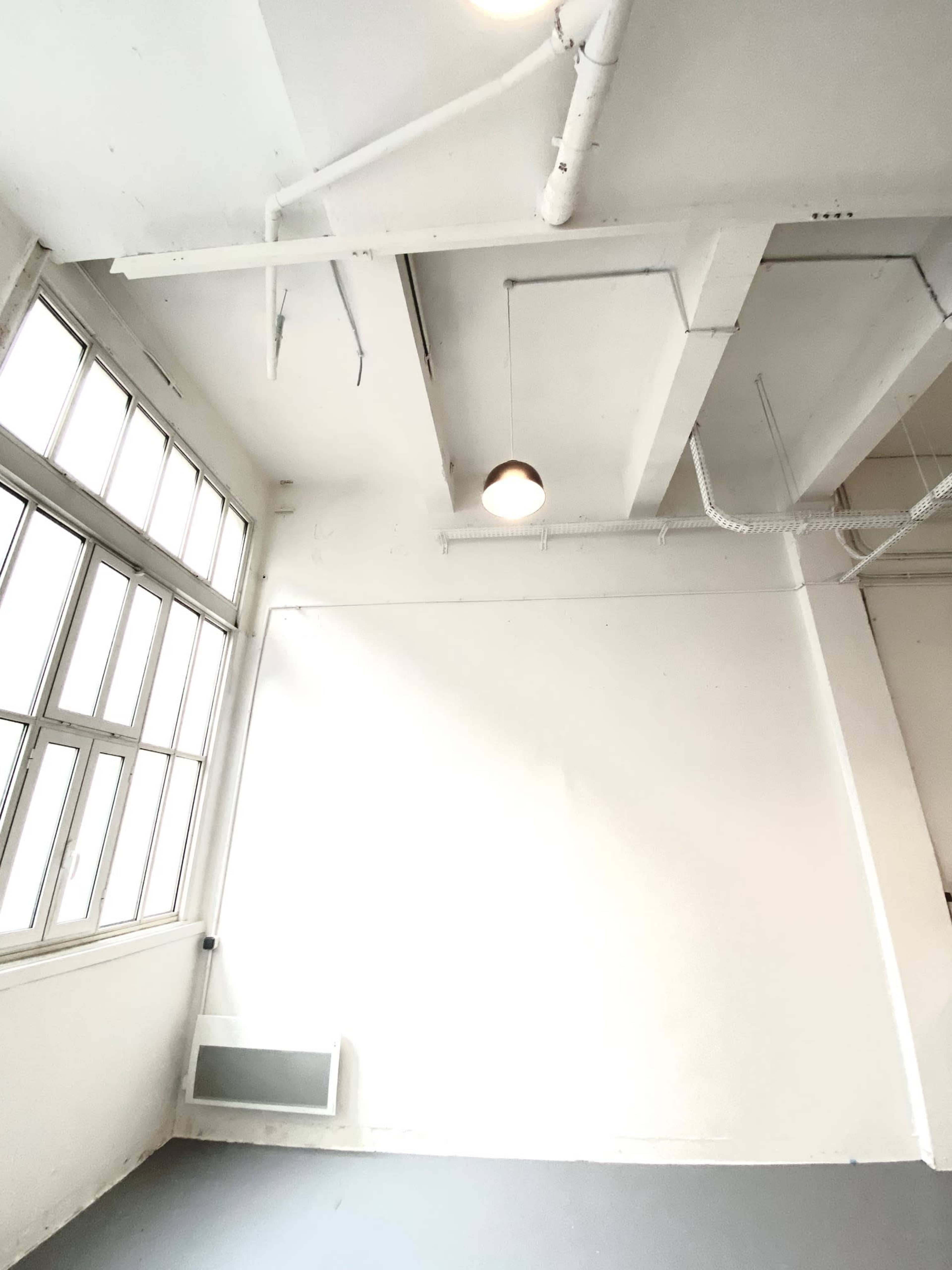 A simple, empty room with large windows, a bare white wall, and a ceiling featuring exposed pipes and a single light fixture.