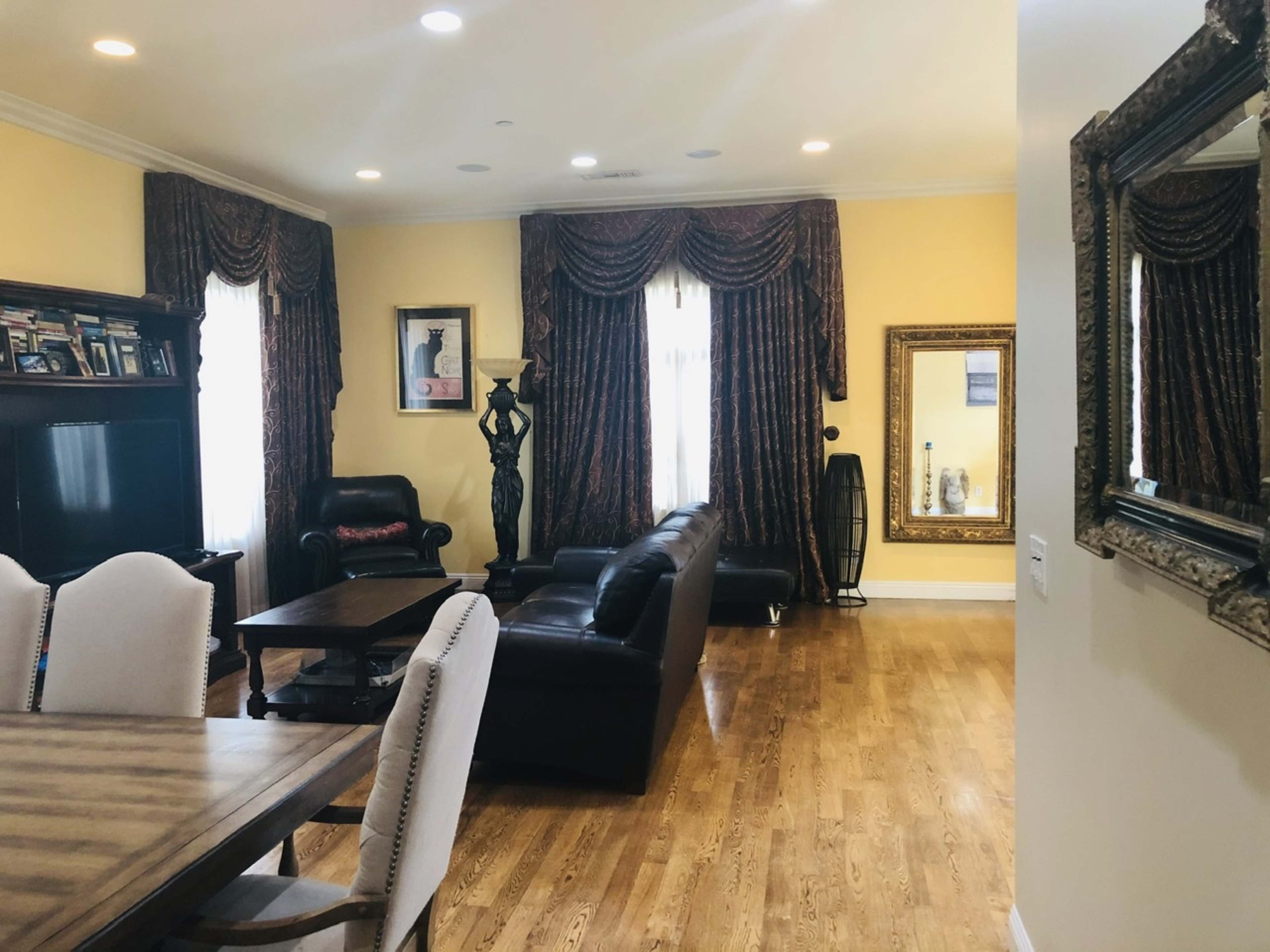 A living room with hardwood floors, a dining table, black leather sofas, intricate curtains, and a large mirror on the wall.