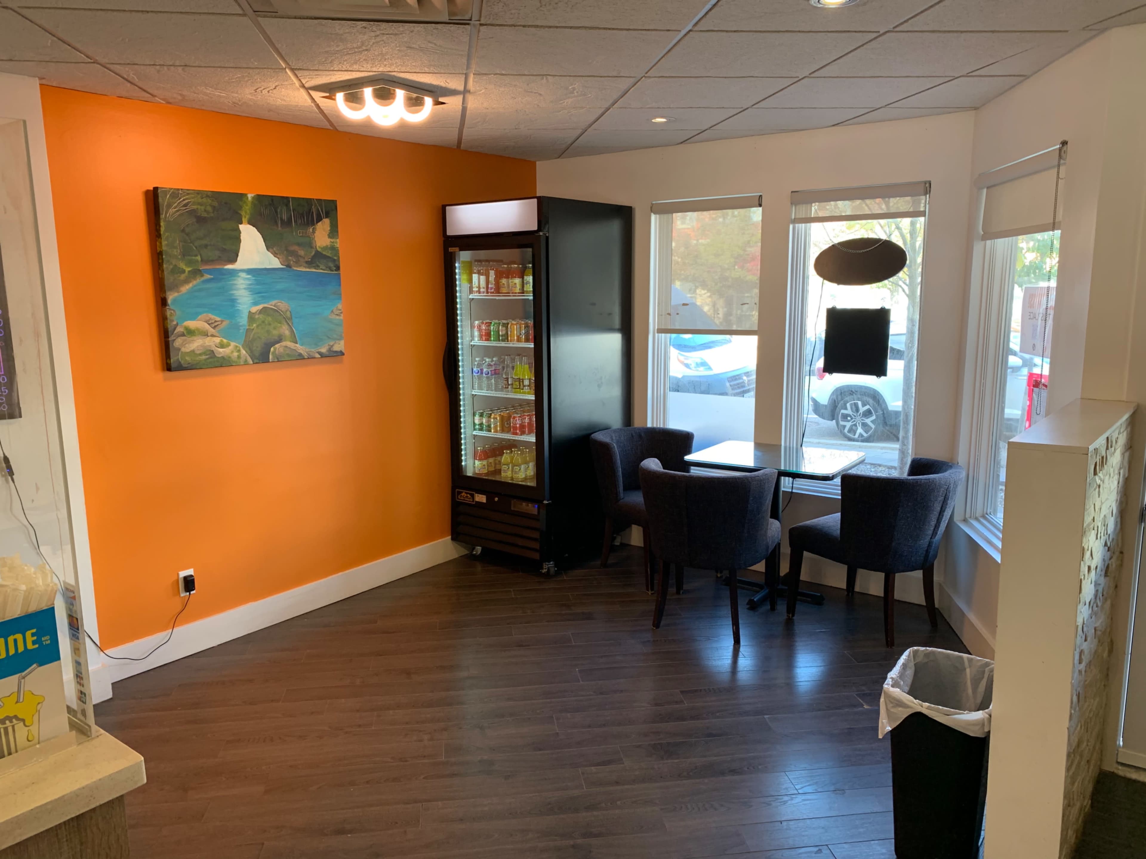 A small café corner with two chairs and a table beside a drink cooler, featuring an orange wall and a landscape painting.