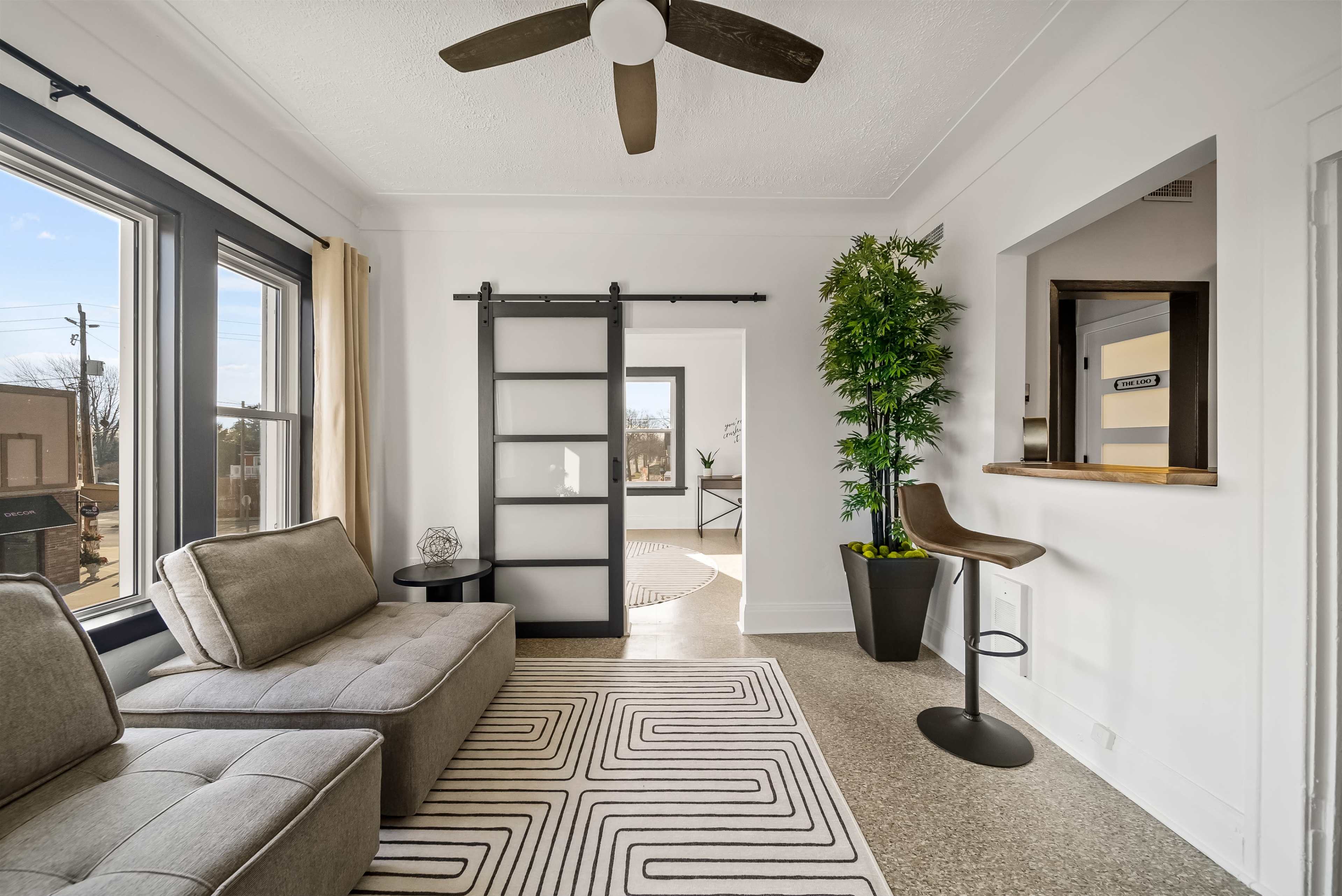 A modern living space with a gray sofa, a sliding black door, a bar stool near a small opening to another room, and a potted plant.