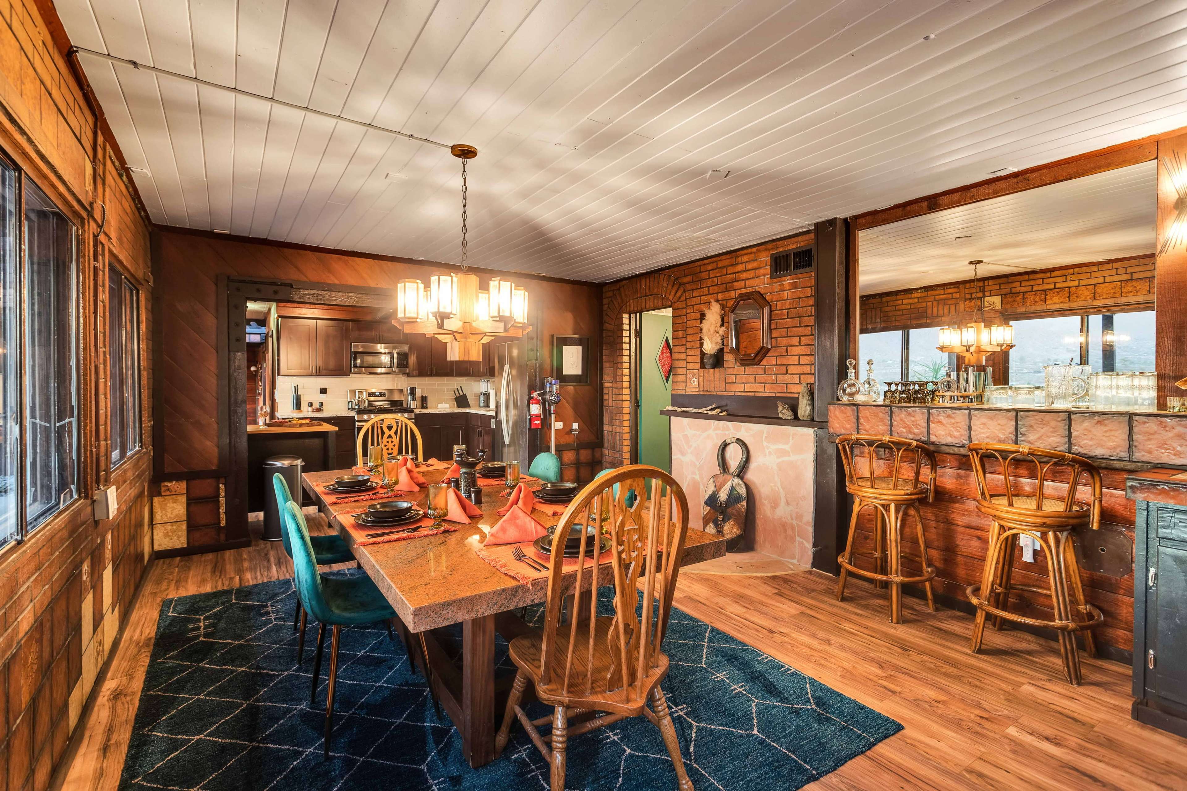 The image shows a rustic dining area with a large wooden table set for a meal, surrounded by wooden chairs, and an adjacent kitchenette with exposed brick walls and a bar area.