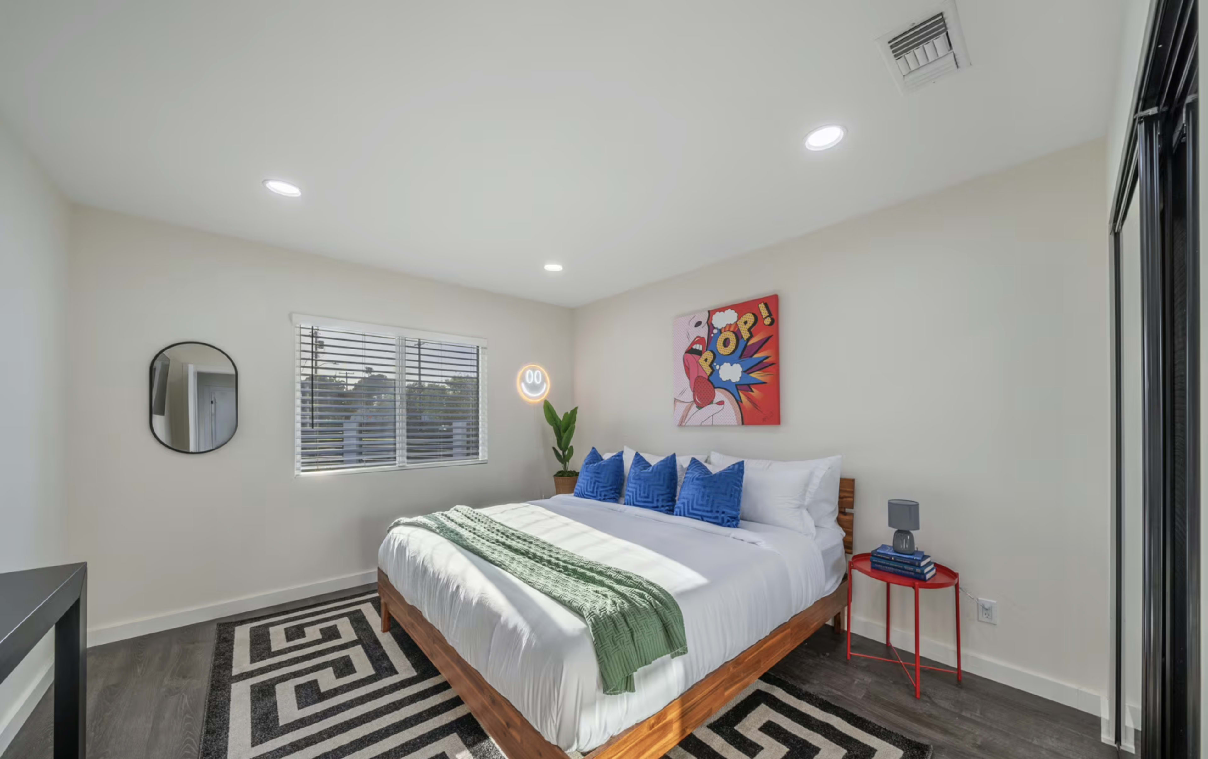 The image shows a modern bedroom with a large bed, decorative pillows, a round mirror, and a colorful wall art piece.