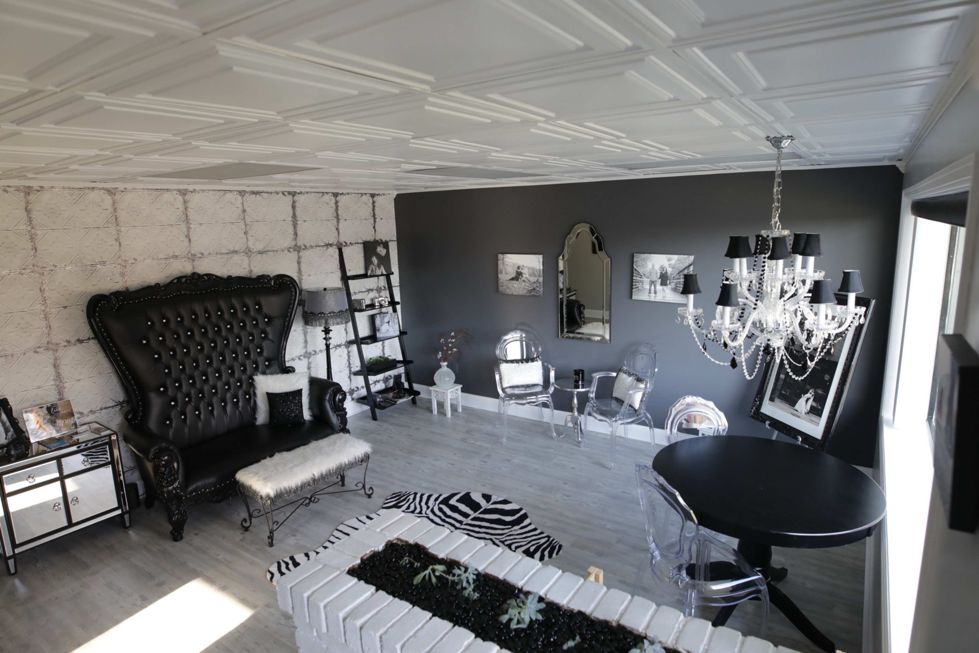 A modern living room featuring a large black tufted sofa, a round black dining table, and a chandelier, with a mix of decorative elements on the walls and floor.