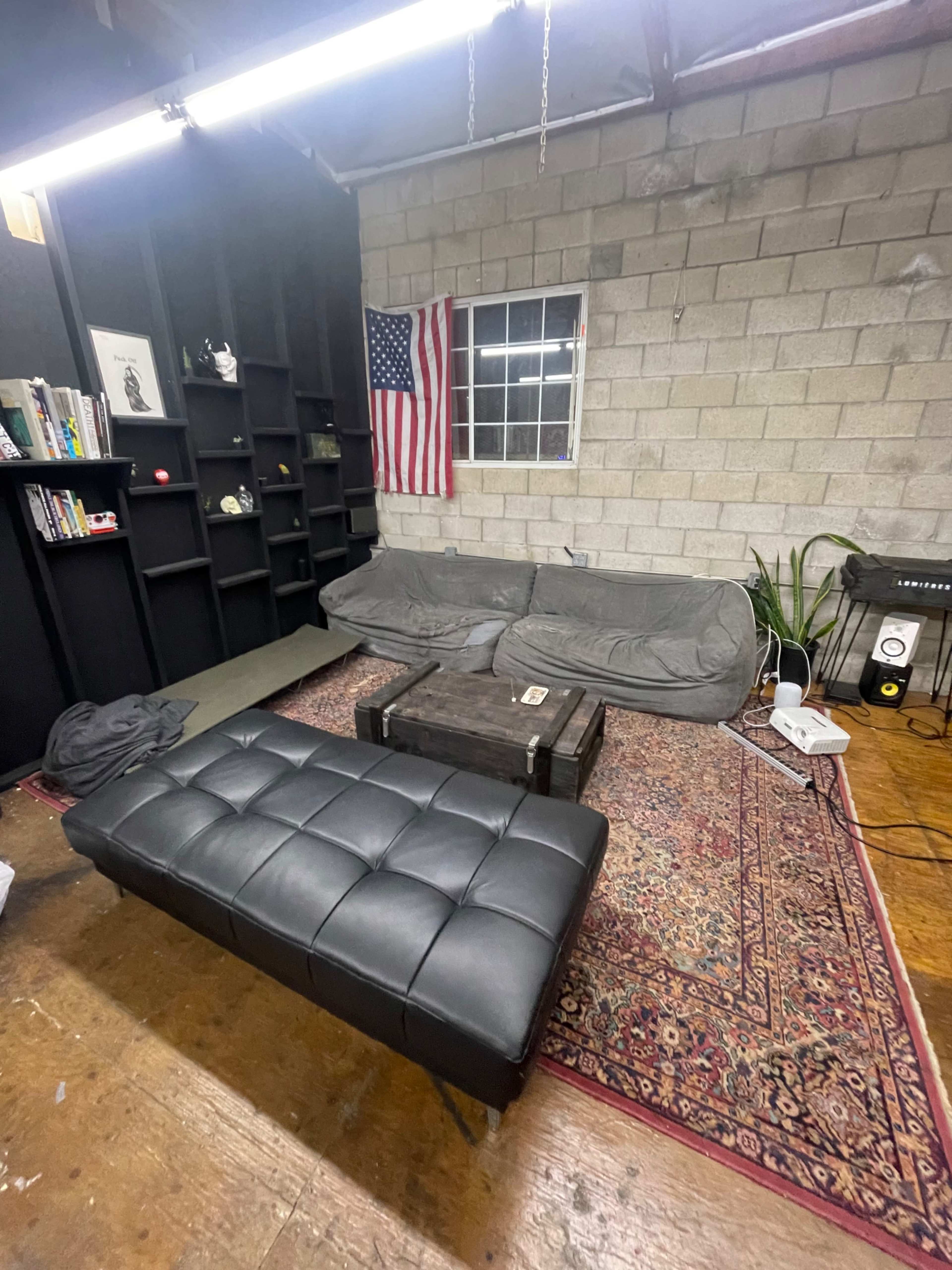 A minimalist living area features a gray sofa and a black ottoman on a patterned rug, with a shelf of books and an American flag on the wall.