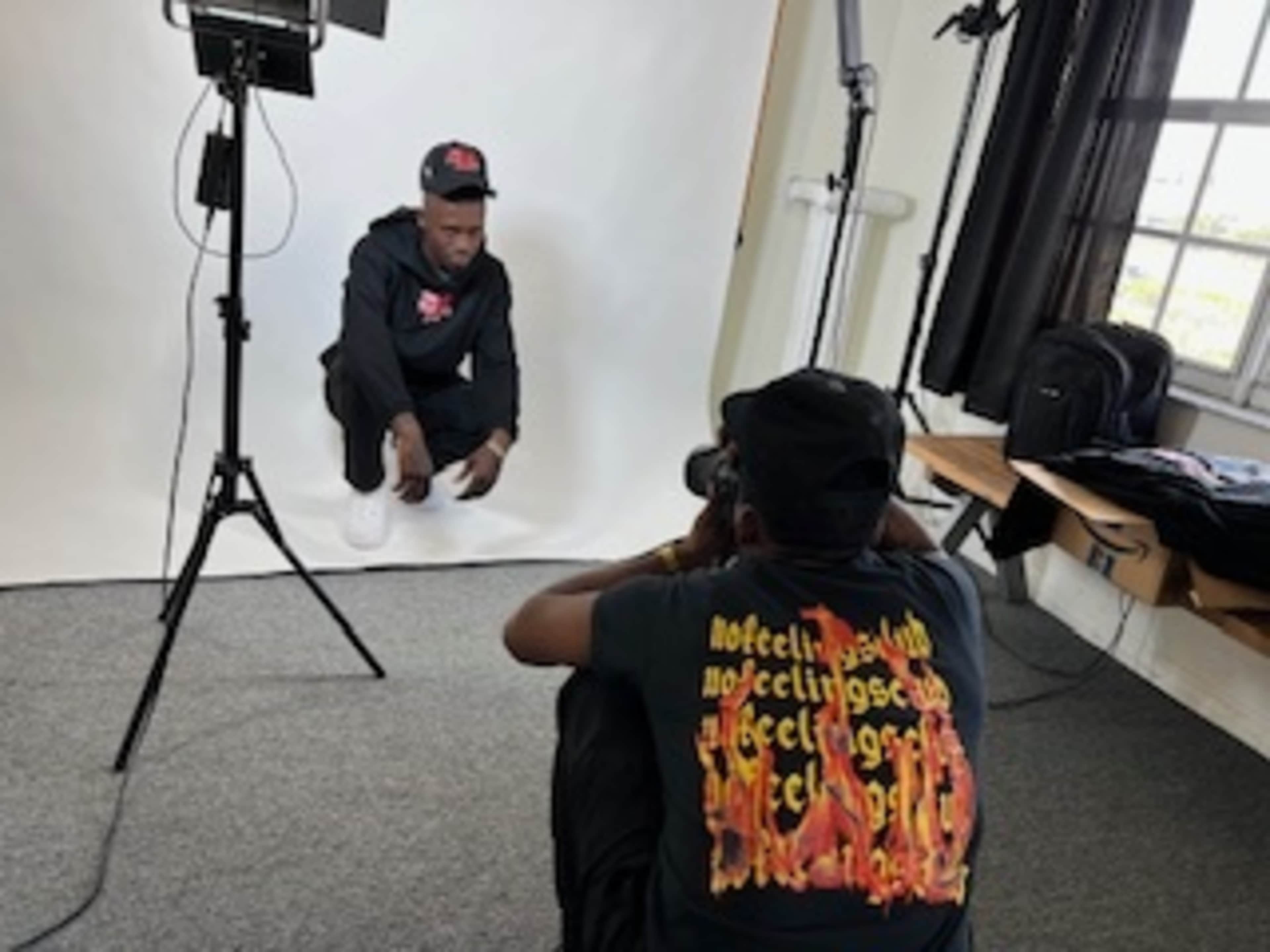 A person sits on the floor in front of a camera setup while another person takes their photo in a studio with a plain backdrop.