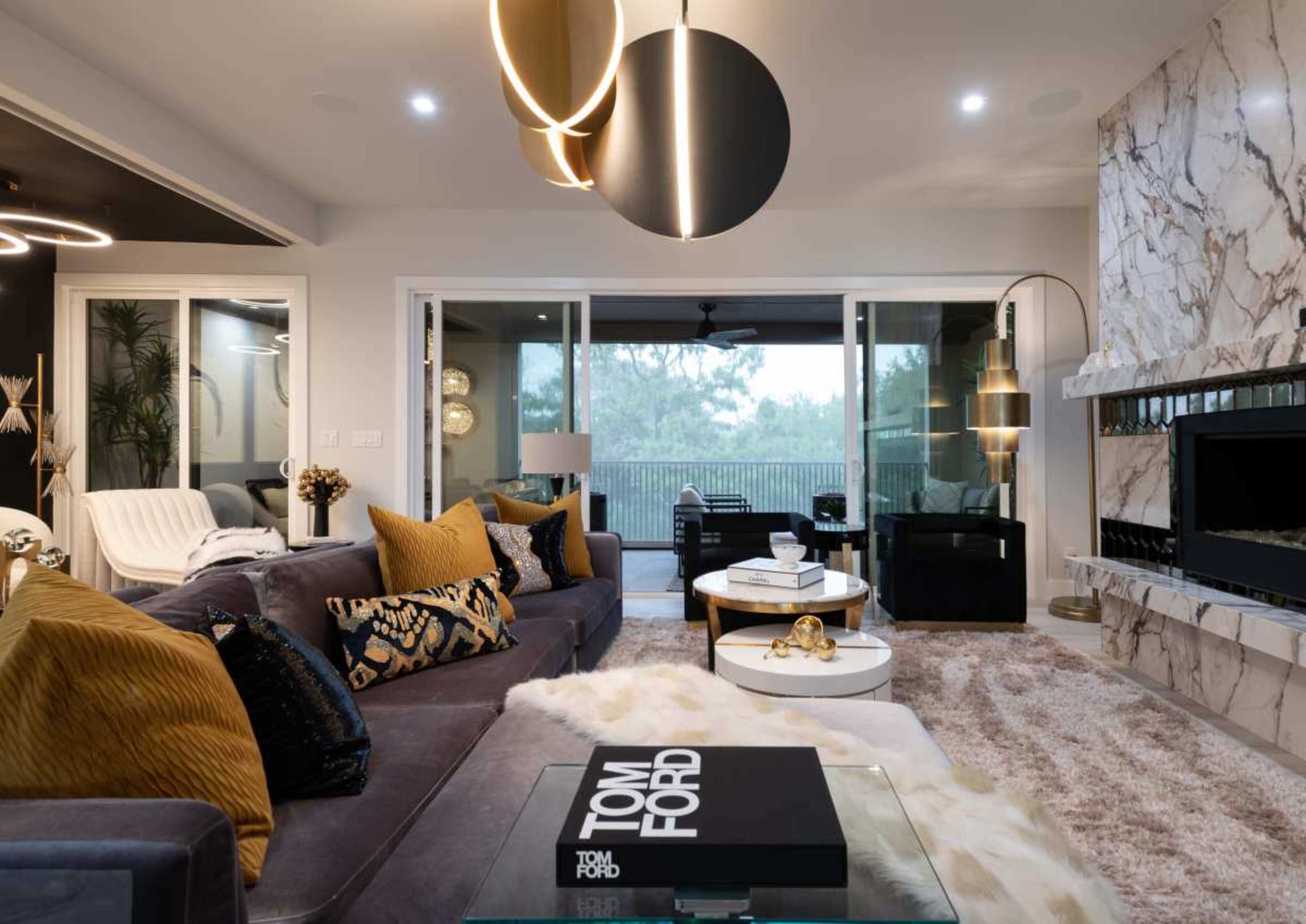 The living room features a modern design with a plush sofa, decorative pillows, a marble fireplace, and large windows offering a view of the outdoors.