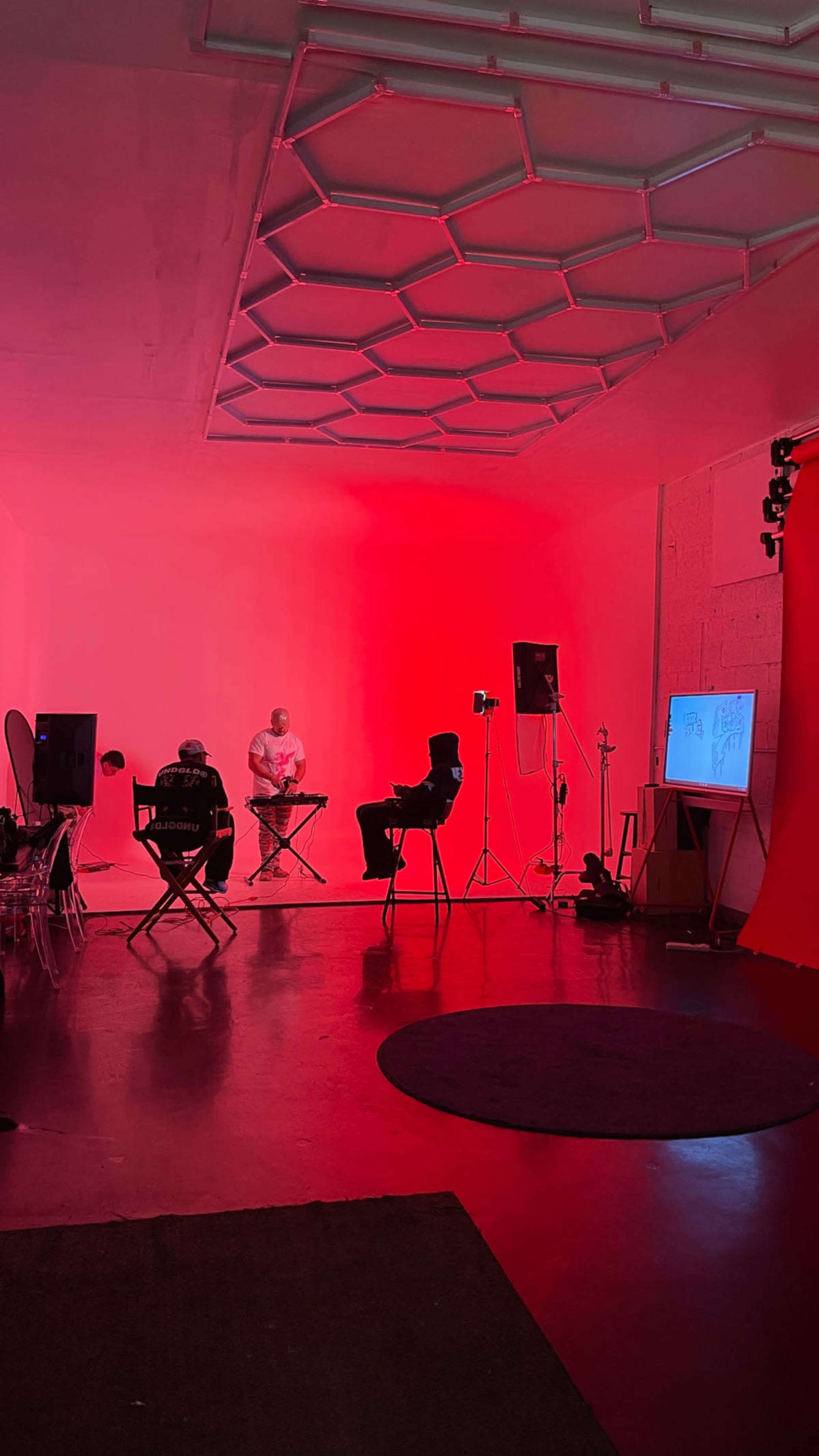 The image shows a modern studio space with a red-lit interior, featuring two seated individuals, a musician playing an instrument, and various equipment set up for recording or performance.