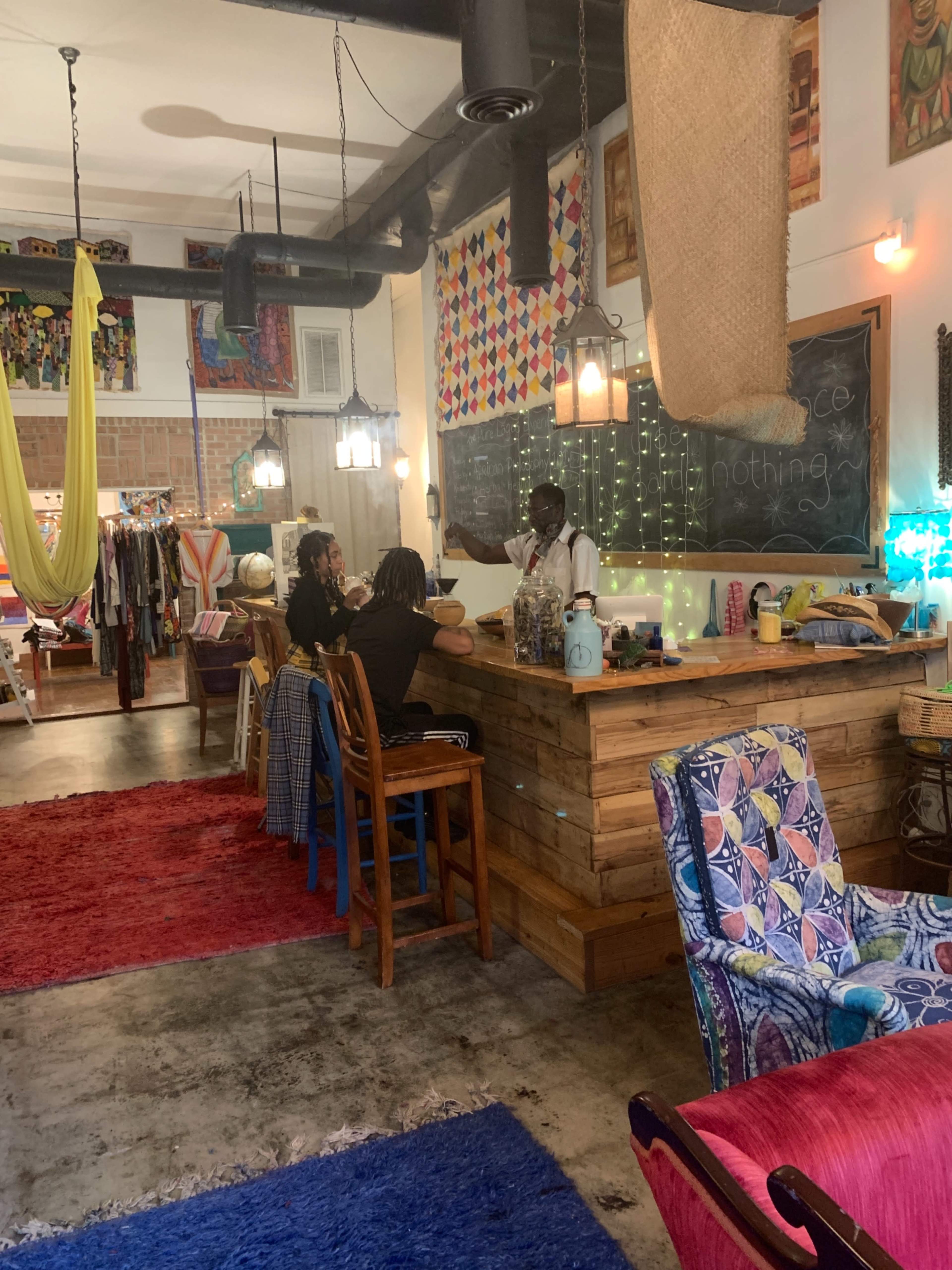 A colorful café interior features a wooden serving counter, with two patrons seated and a server behind the counter, surrounded by vibrant textiles and art.