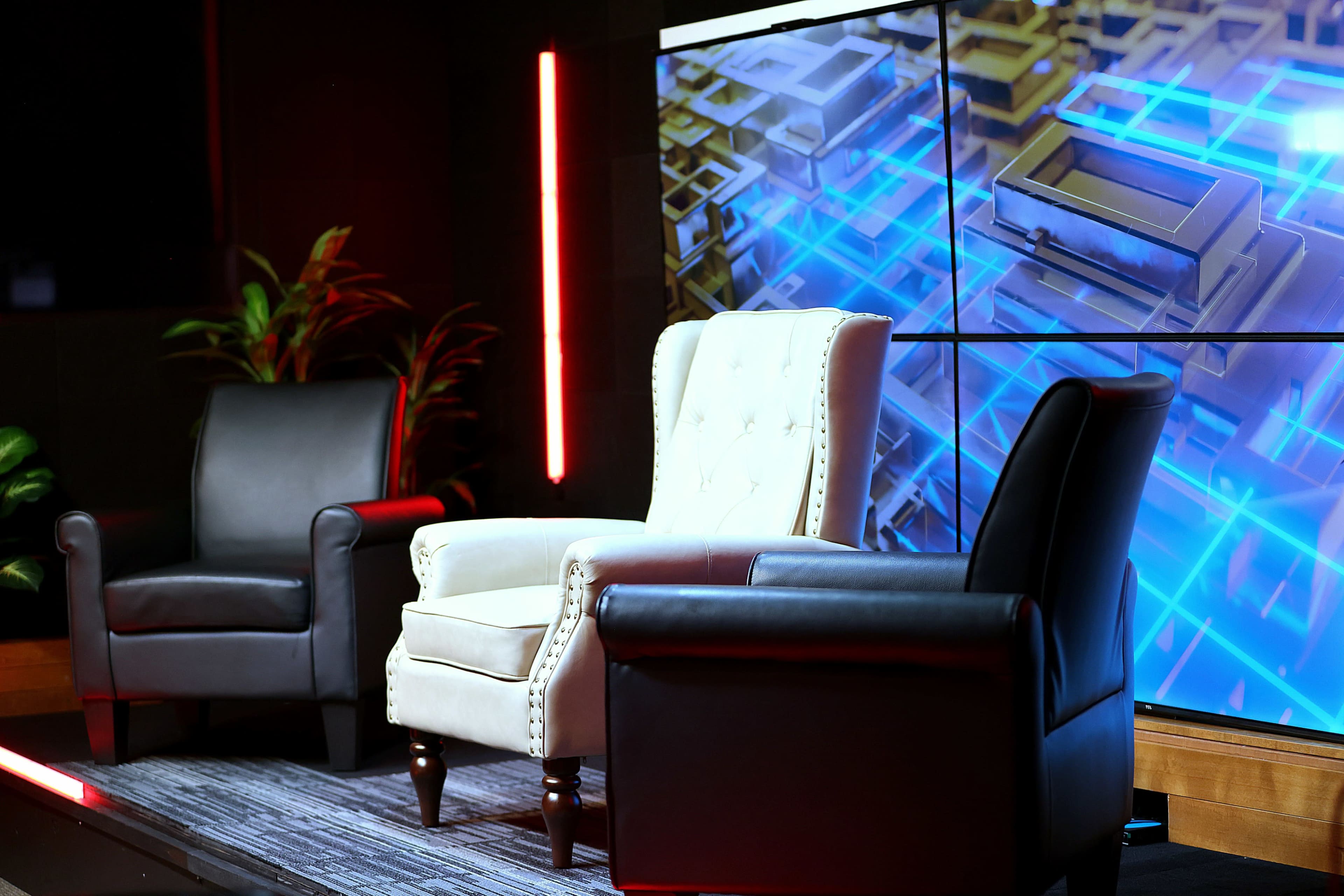 The image shows three armchairs, two black and one white, arranged on a stage with a large digital display behind them.