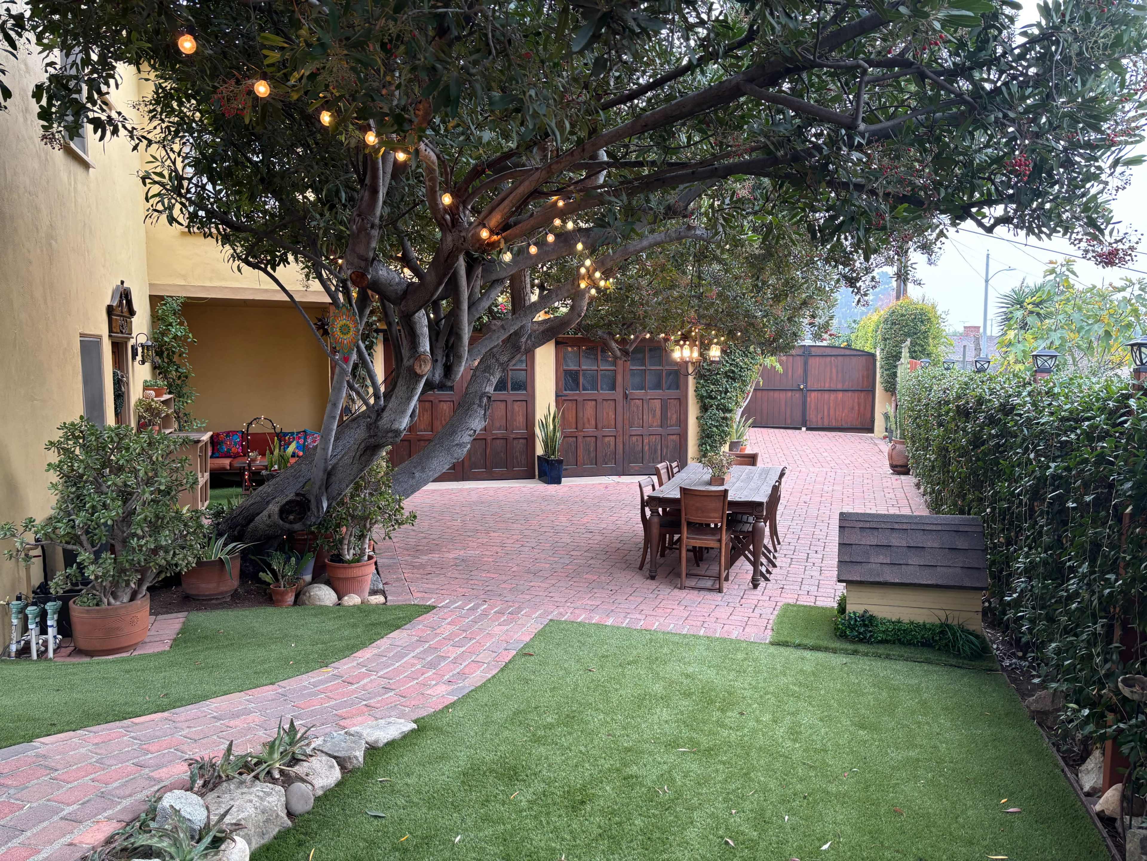 A brick-paved courtyard features a large tree with string lights, surrounded by potted plants and wooden furniture.