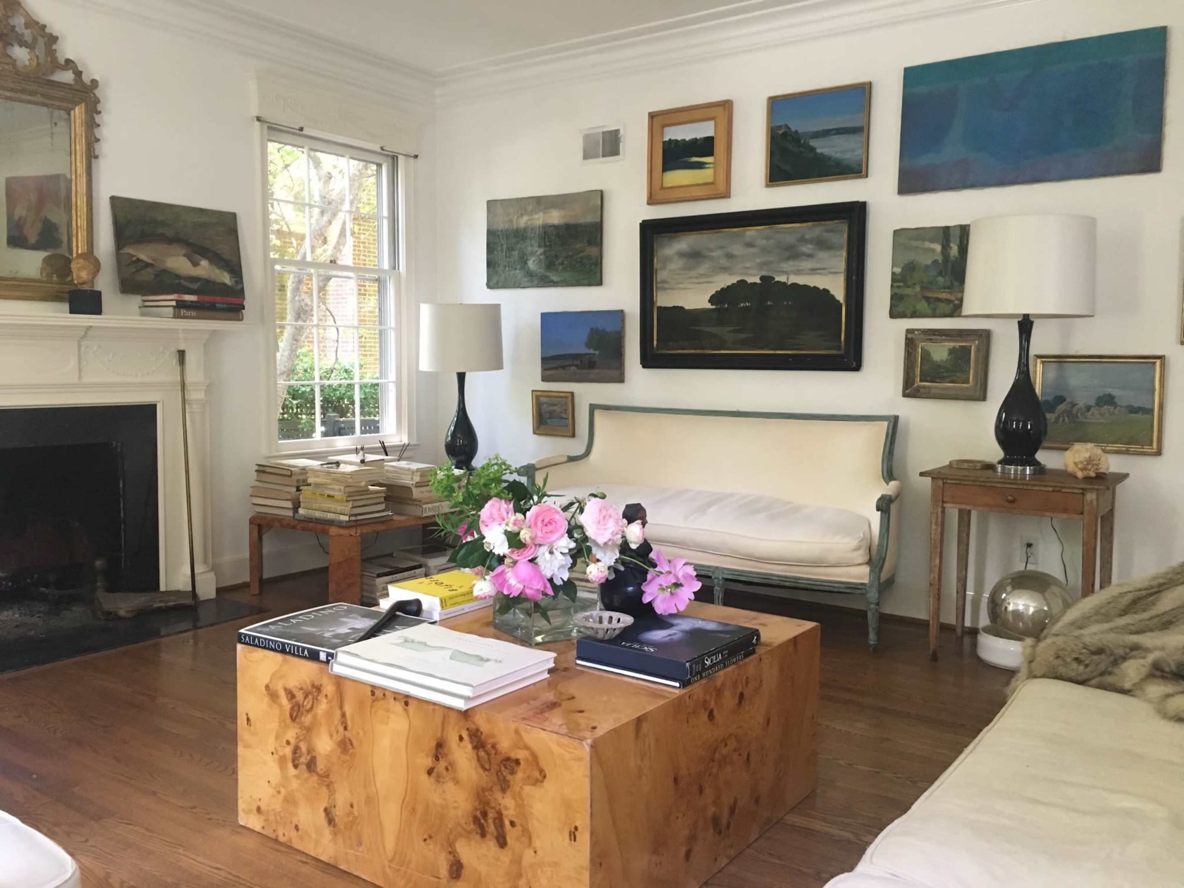 A living room features a coffee table made of wood, a white couch, and a wall adorned with various framed paintings, along with large lamps on either side.