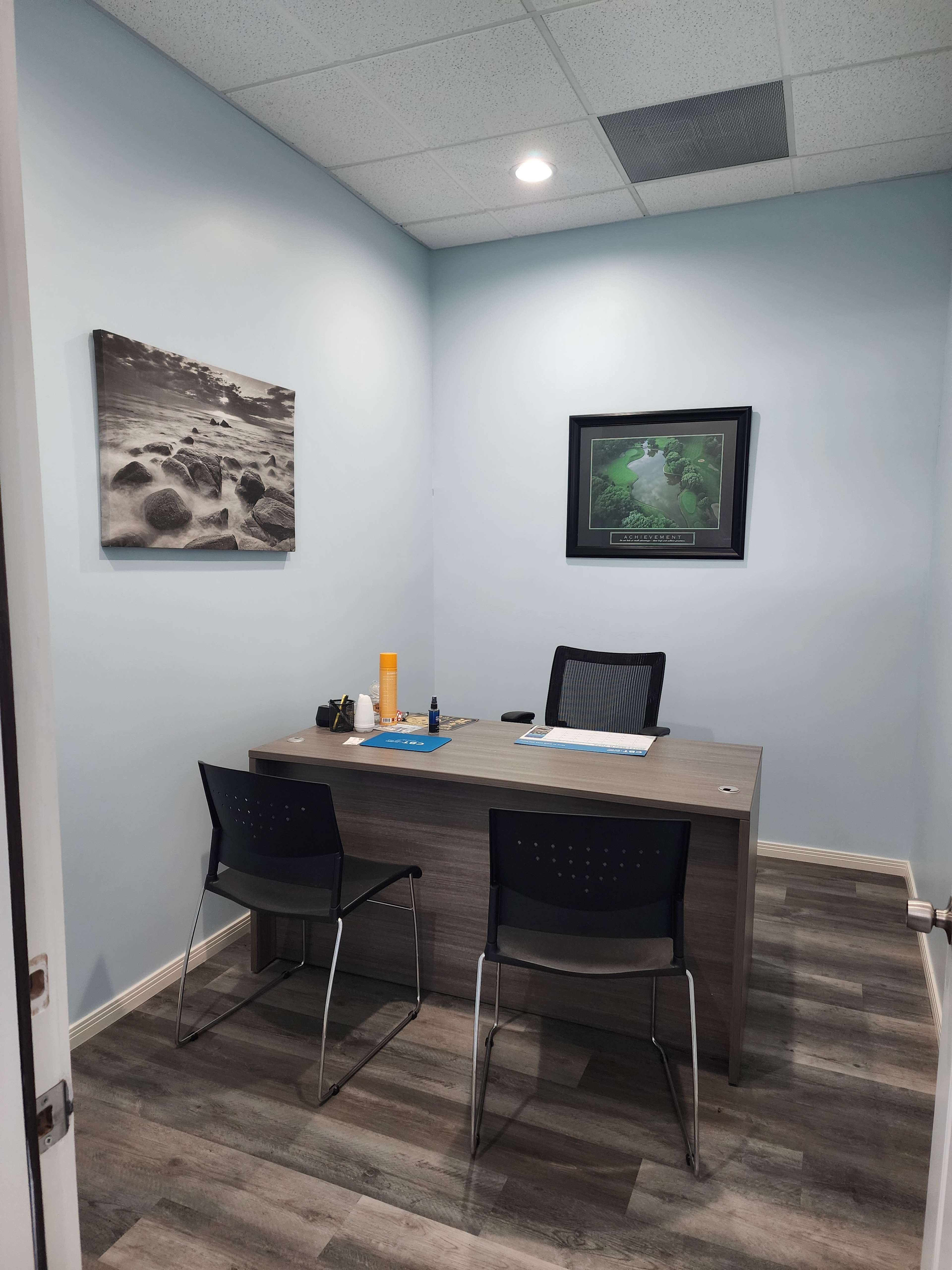 The image shows a small office space with a desk, two chairs, and framed photographs on the walls.