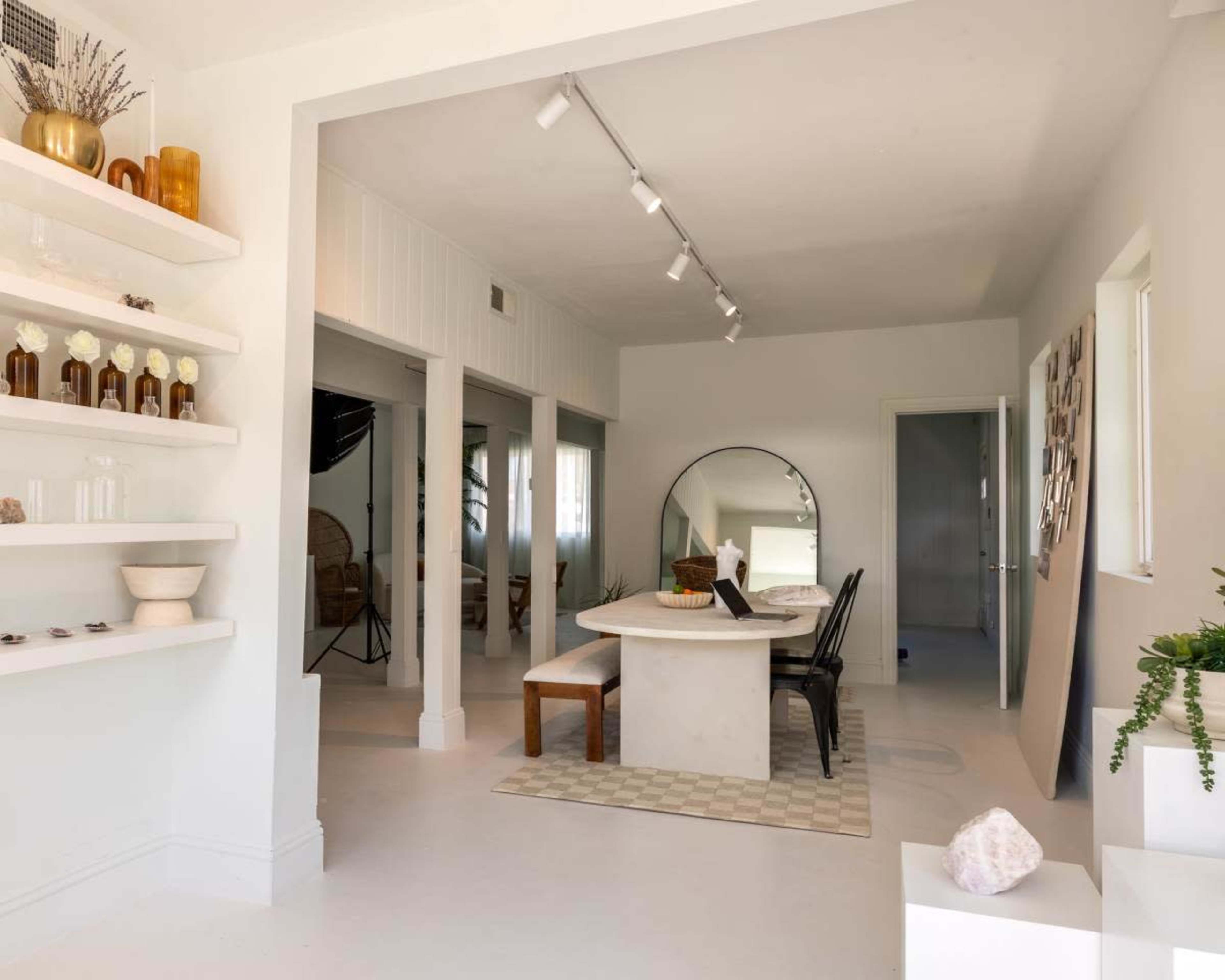 The image shows a bright, modern interior with a dining table surrounded by black chairs, a large mirror on the wall, and shelves displaying decorative bottles and plants.