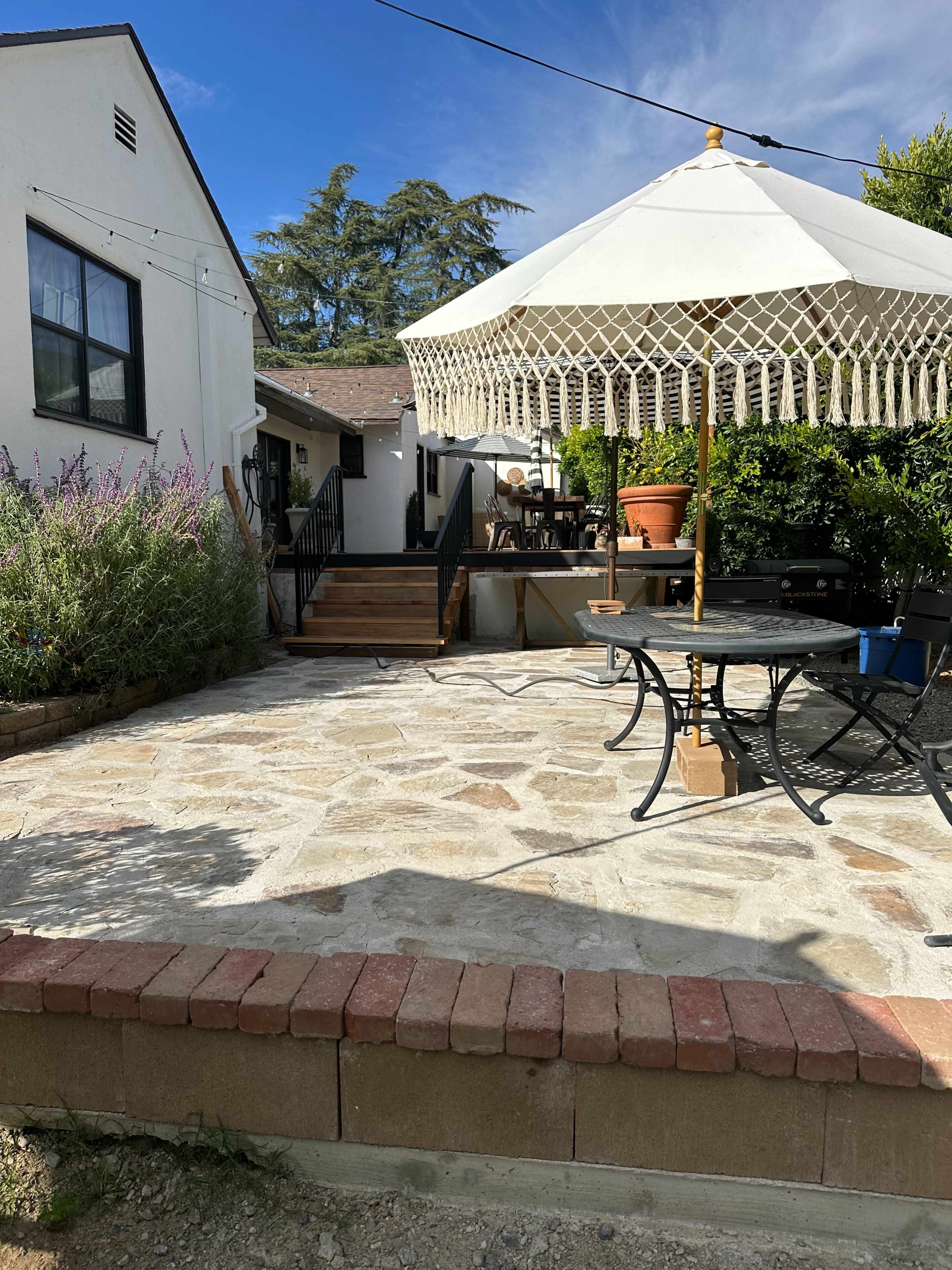 The image shows a stone patio area with a circular table and chairs beneath a large umbrella, bordered by plants and connected to a building.