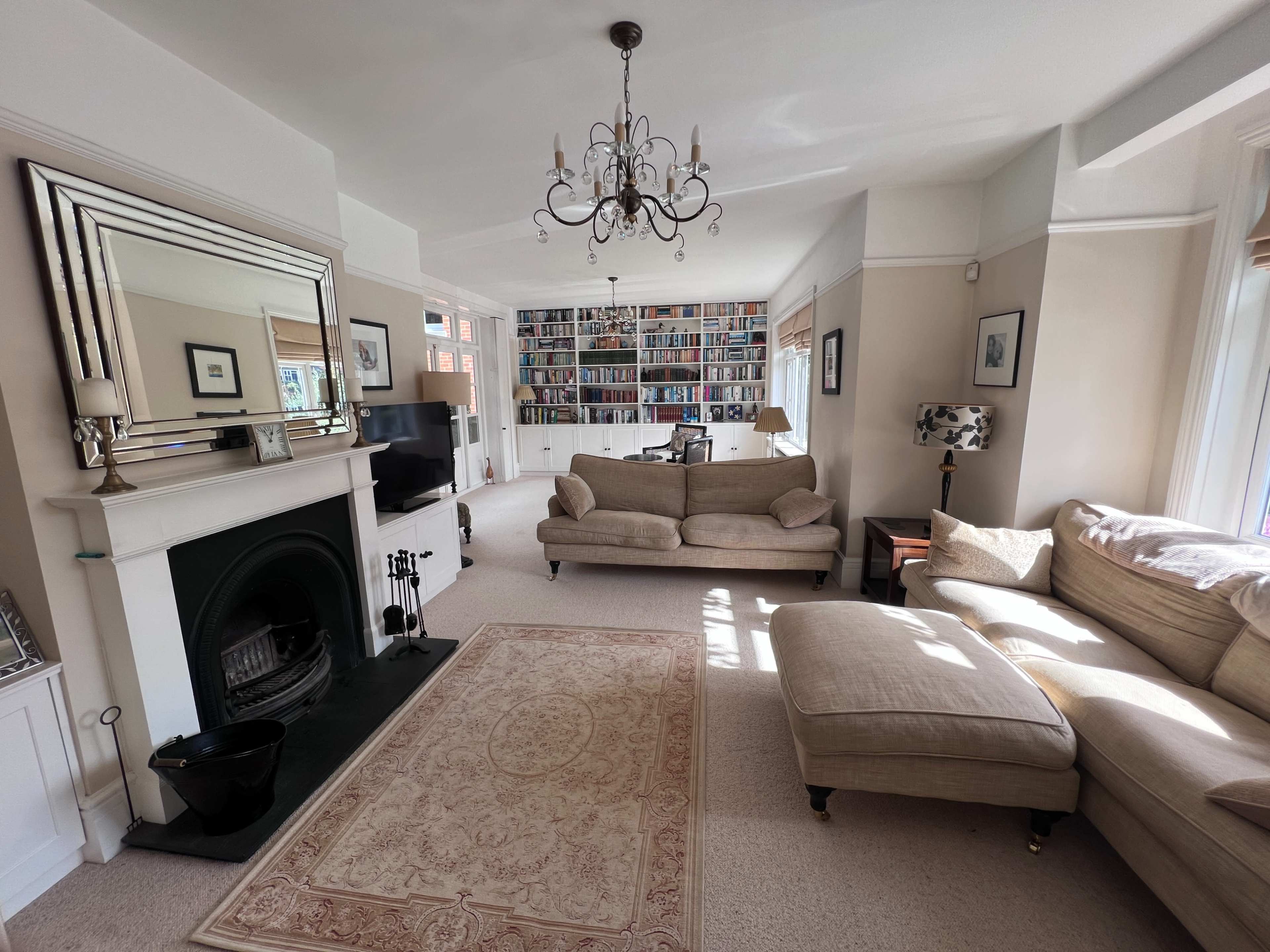 A spacious living room features a large bookshelf filled with books, a chandelier, a fireplace, and multiple seating areas.