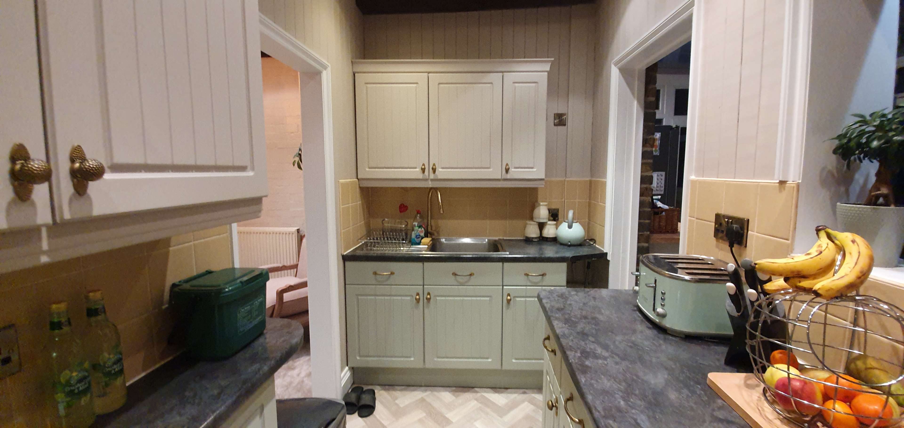 A modern kitchen with light-colored cabinets, a stainless steel sink, and a counter filled with a fruit basket and kitchen appliances.
