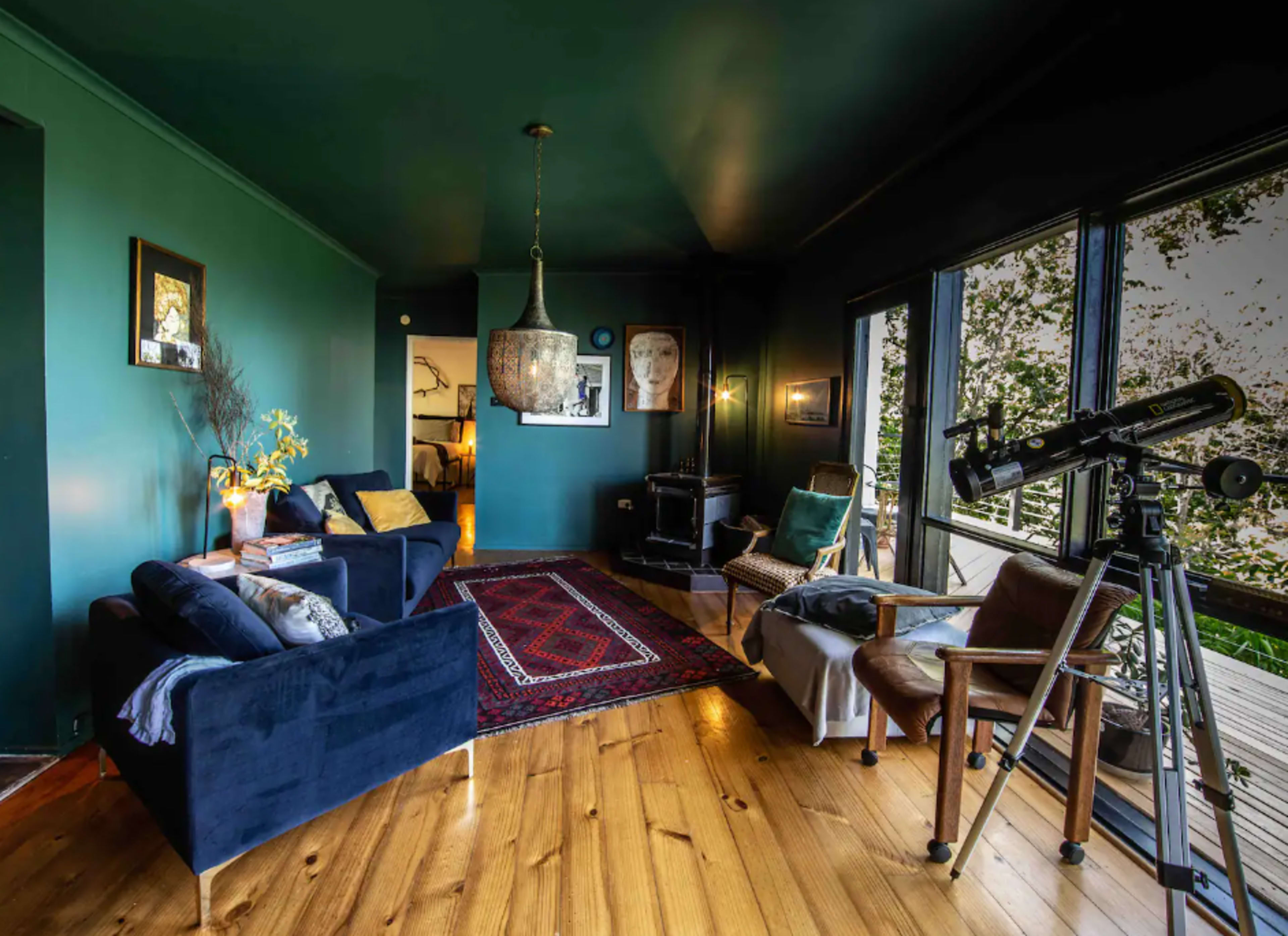 A cozy living room features dark green walls, wooden flooring, a patterned rug, a telescope, and large windows that look out onto nature.