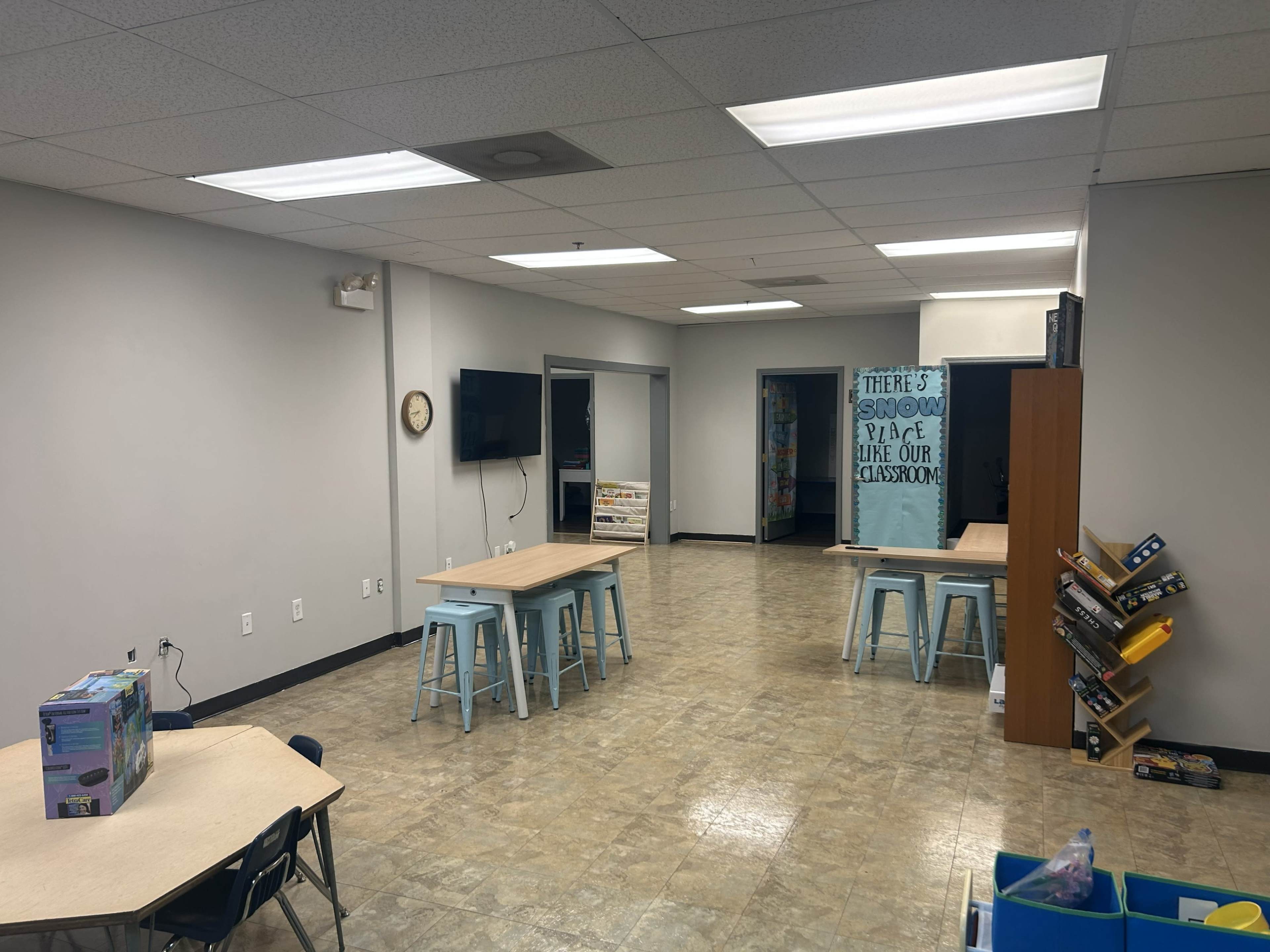 A classroom is shown with several tables, blue stools, and shelves filled with books against the walls.