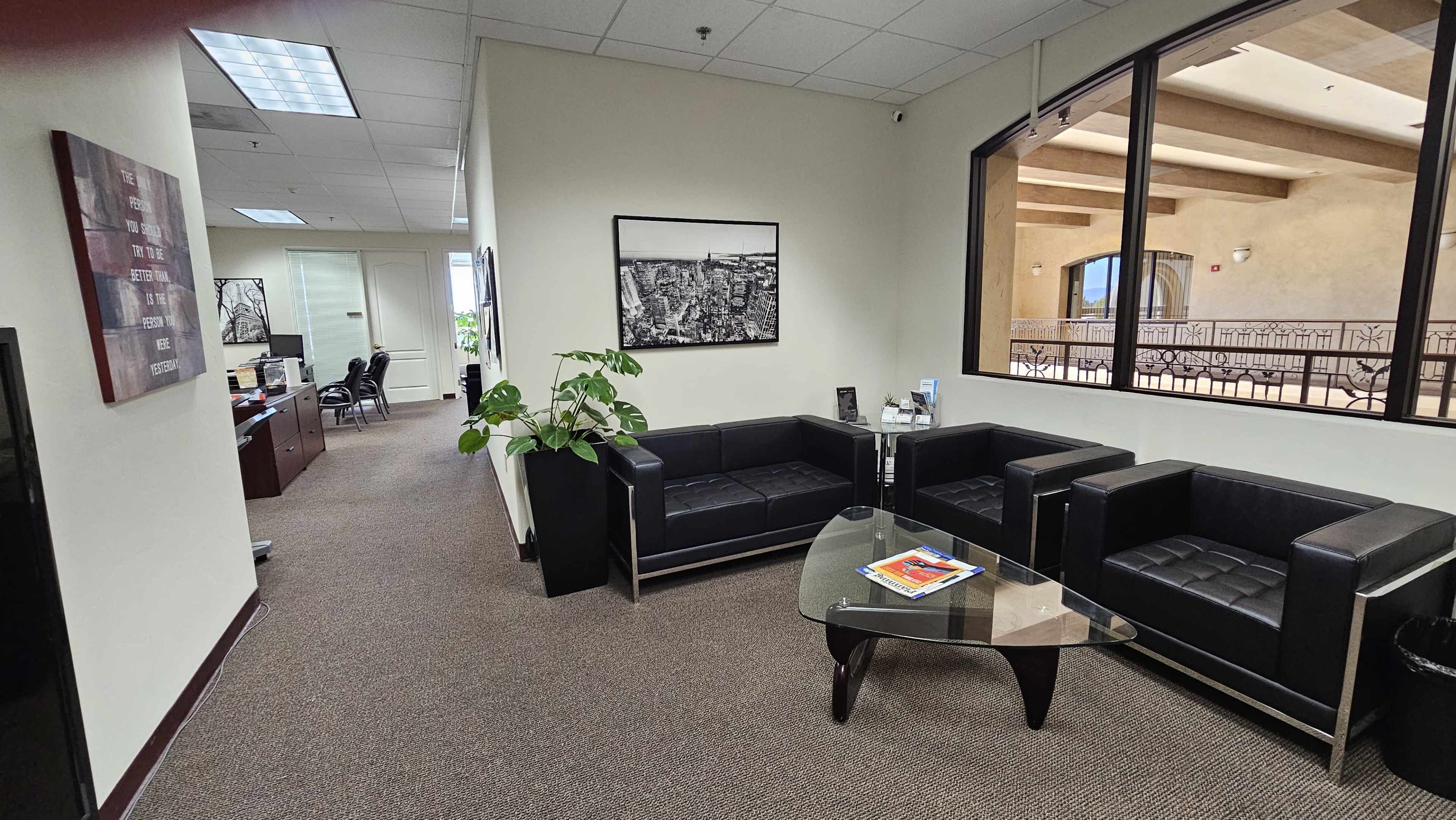 A modern office waiting area features black leather couches, a glass coffee table, and a plant, with an open room leading to a desk and office space.