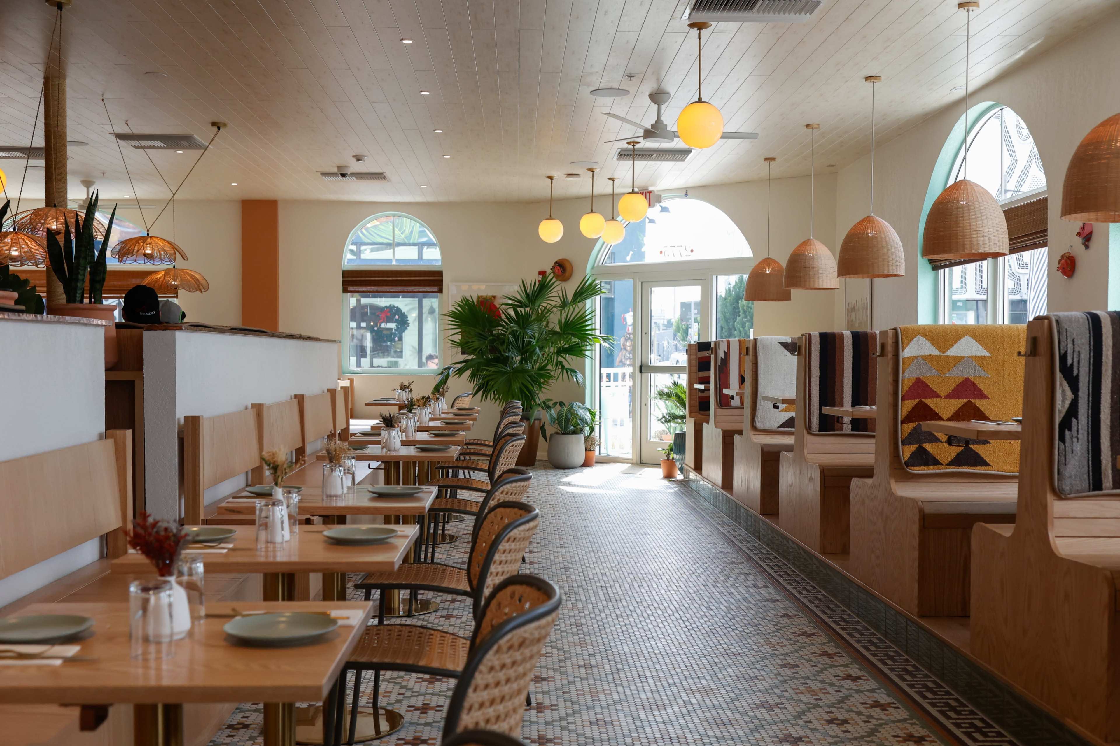 The image shows a brightly lit restaurant interior with wooden tables, wicker chairs, and decorative booths, featuring large windows and pendant lighting.