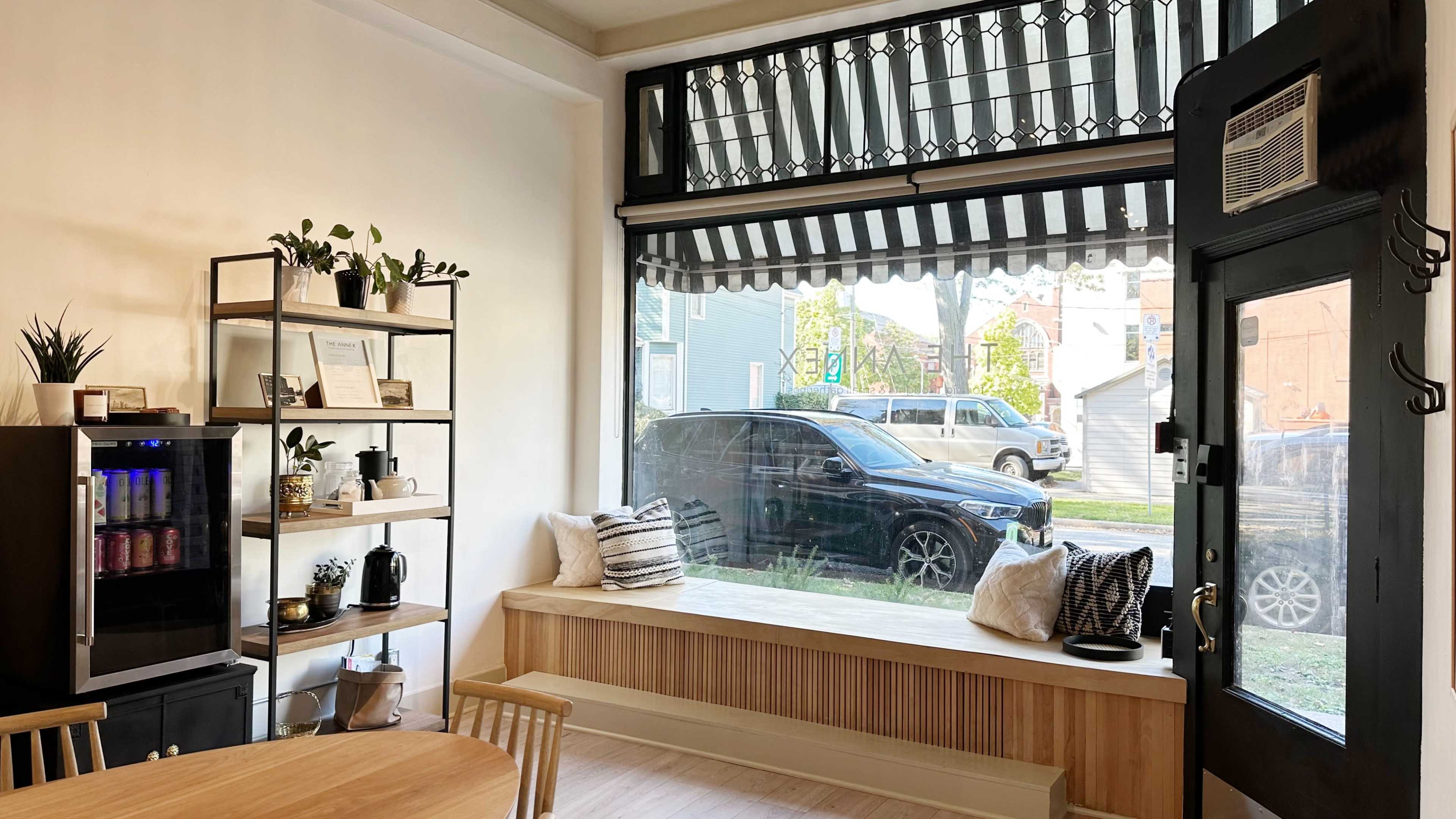 The image shows a cozy interior space featuring a wooden bench by a window, a dining table with chairs, a shelving unit with plants, and a view of a parked car outside.