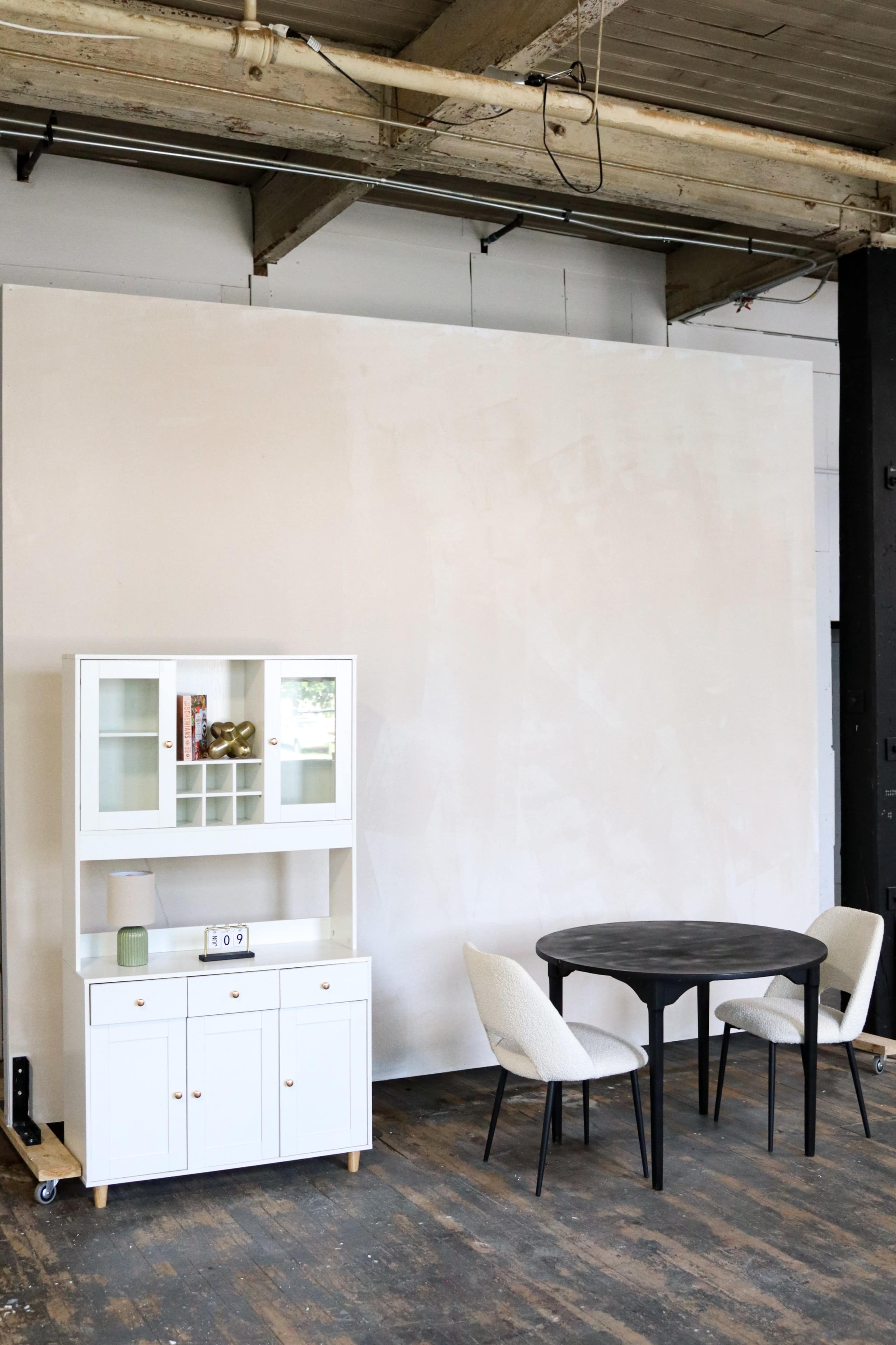 A white cabinet and a round black table with two chairs are positioned against a beige wall in a spacious room with wooden flooring.