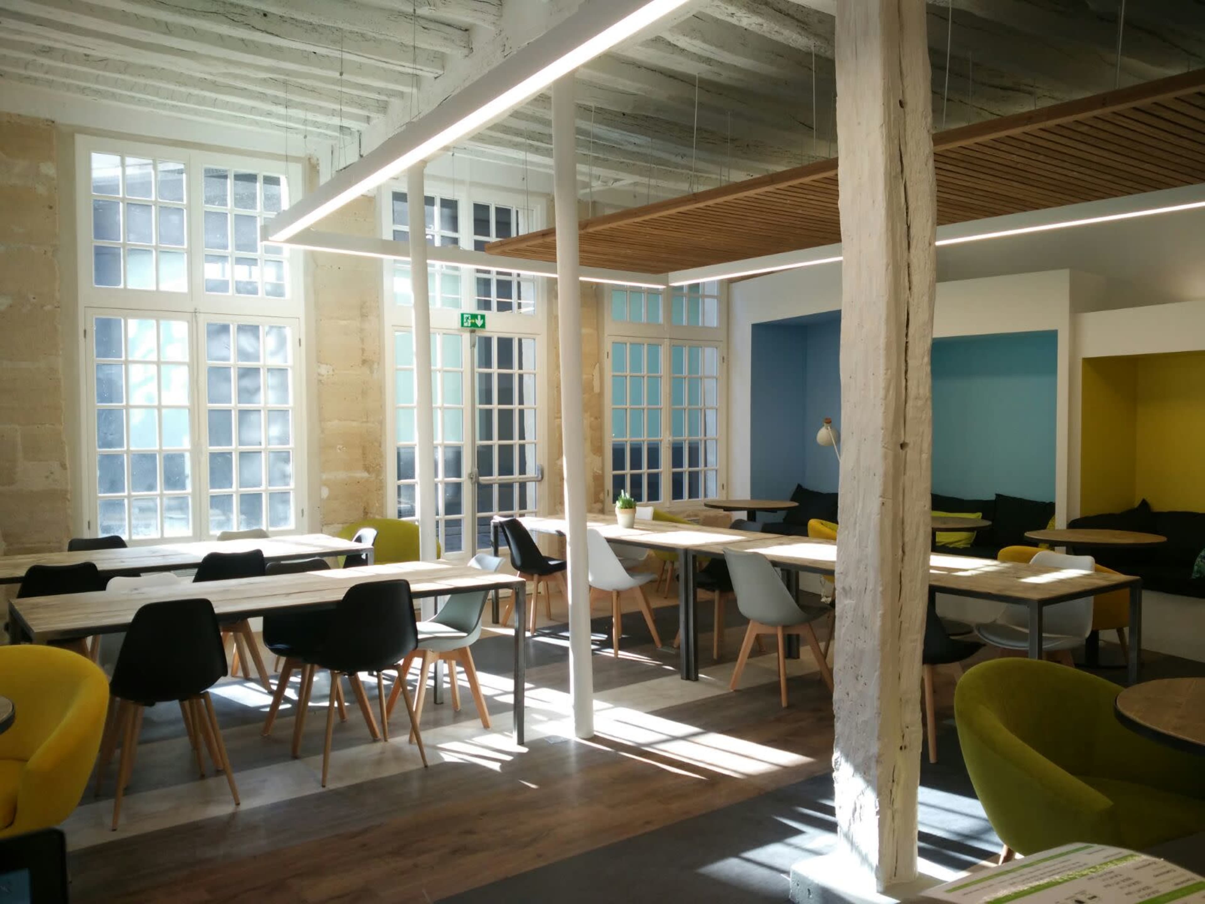 A bright, modern workspace features wooden tables and colorful chairs arranged around large windows letting in natural light.