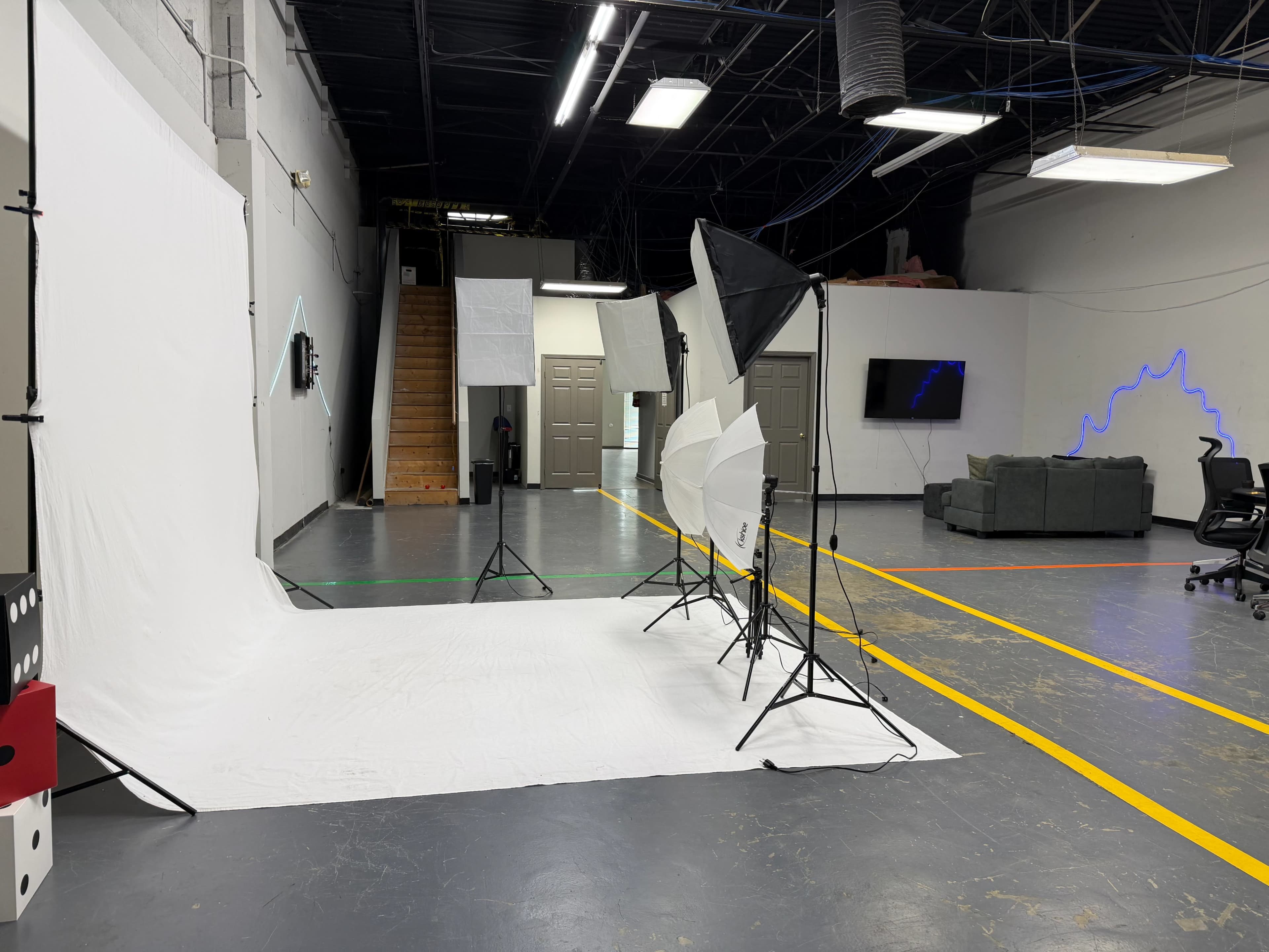 The image shows a photography studio with a white backdrop, multiple light stands, and a seating area featuring a couch and a television.